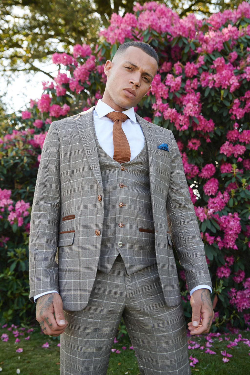 A man wears the marcdarcy RAY - Tan Check Blazer with a white shirt and brown tie, standing confidently outdoors before pink flowering bushes, his hands relaxed by his sides.