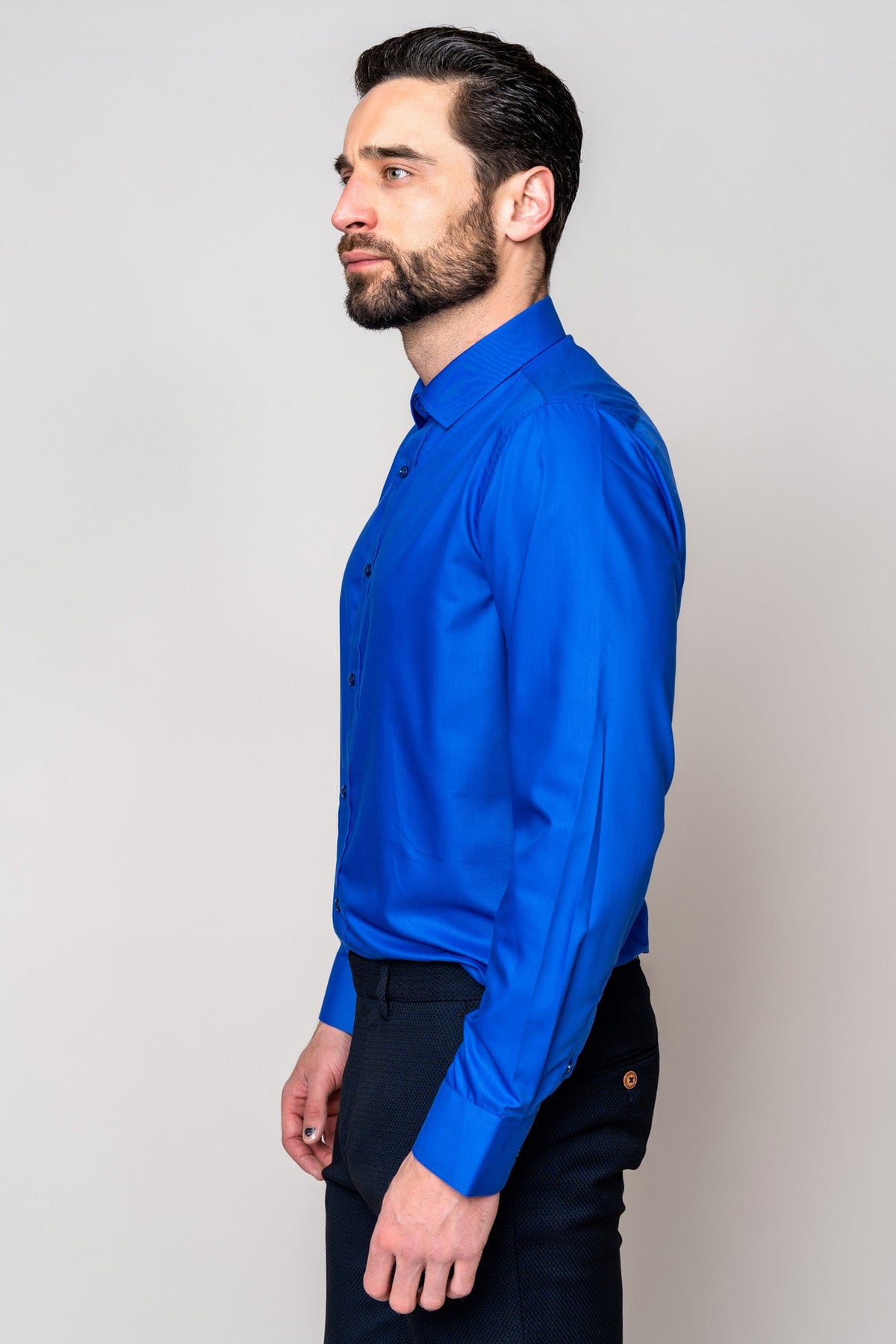 A man with dark hair and a beard stands in profile, wearing the Marc Darcy CARTER - Royal Long Sleeve Shirt with dark pants against a plain light background.