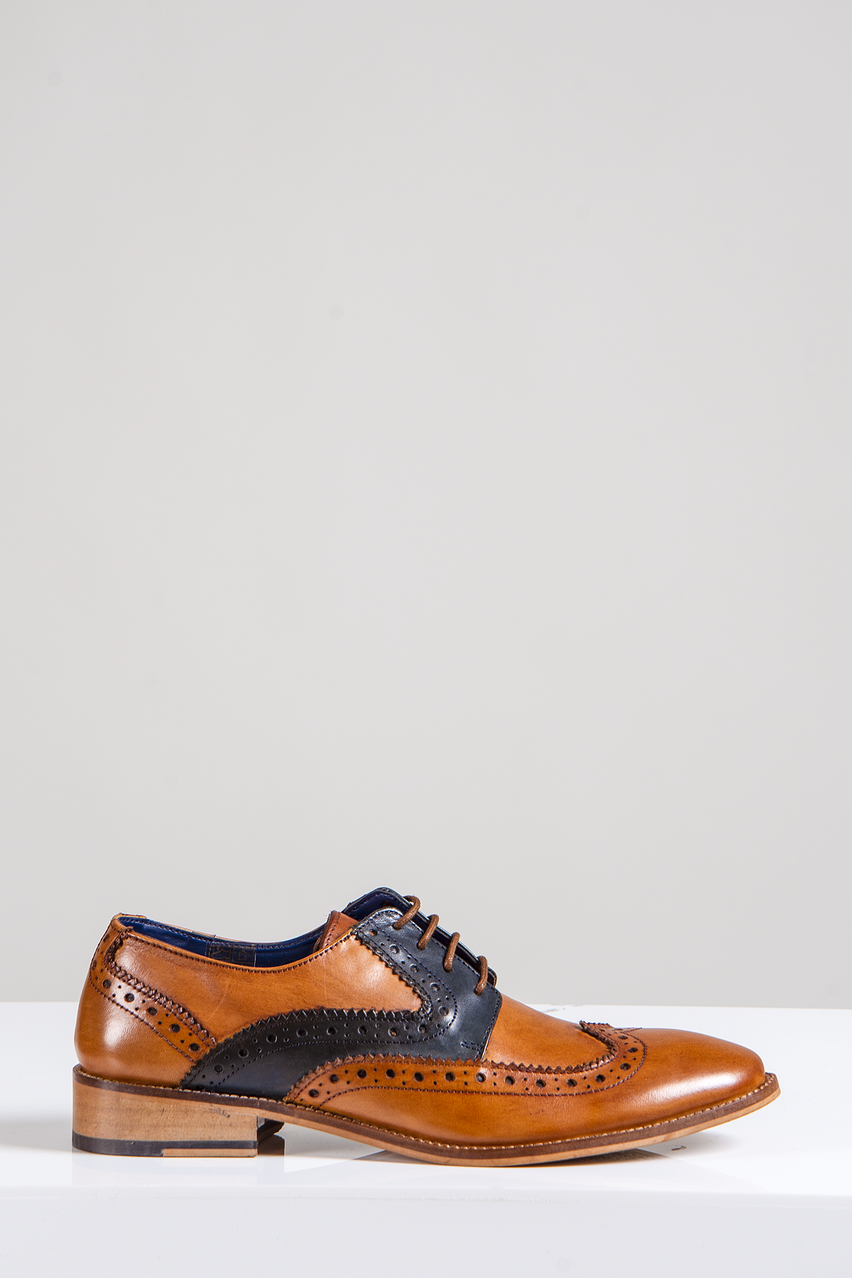 The Marc Darcy RILEY is a tan and navy leather contrast Oxford brogue shoe with decorative perforations, hand-stitched detail, dark laces, and a low wooden heel, displayed on a white surface against a plain light background.
