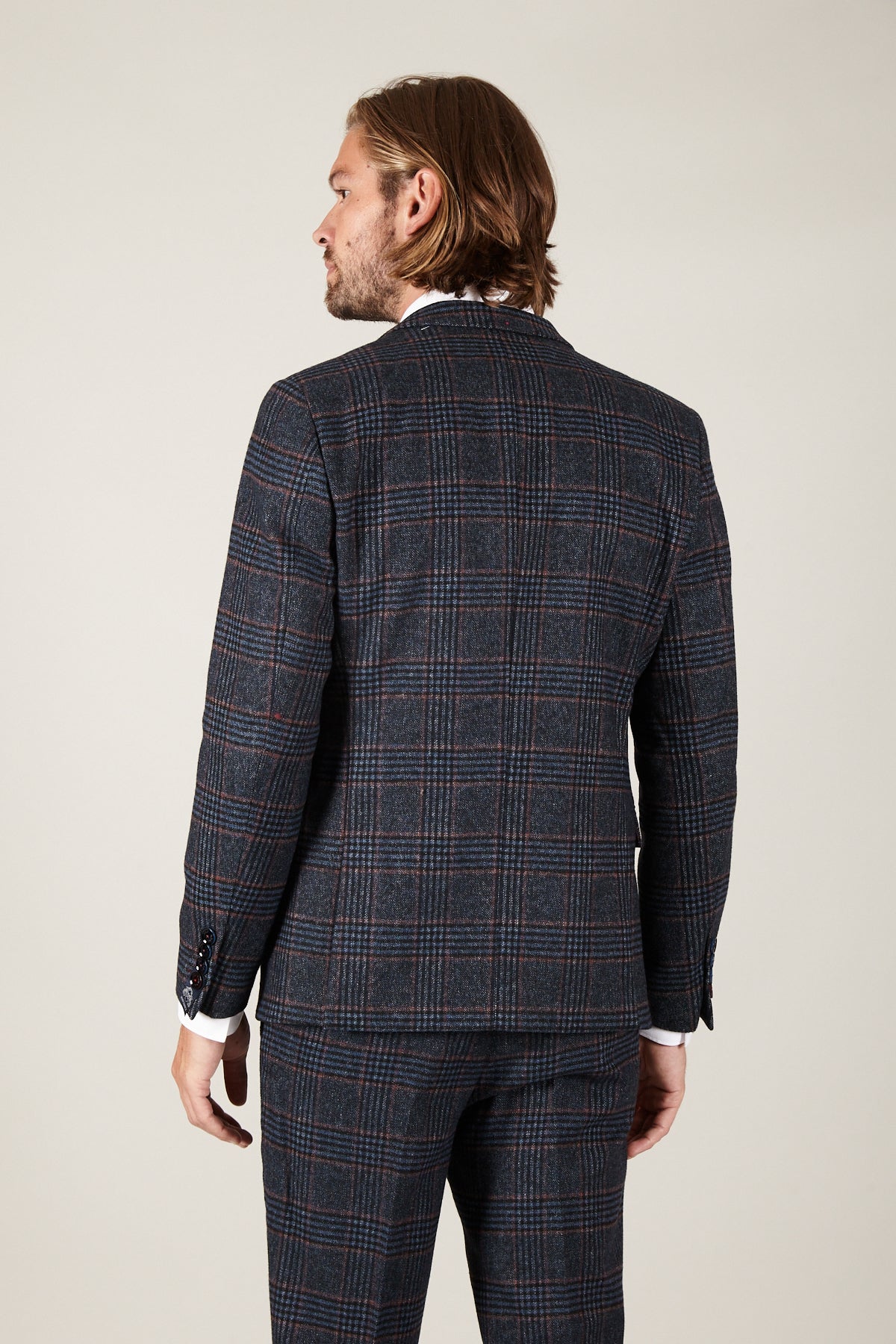 A man with shoulder-length hair is seen from the back in the marcdarcy LUCA Navy Check Tweed Blazer, ideal for vintage lovers. He stands against a plain light background, his left arm slightly bent at his side.