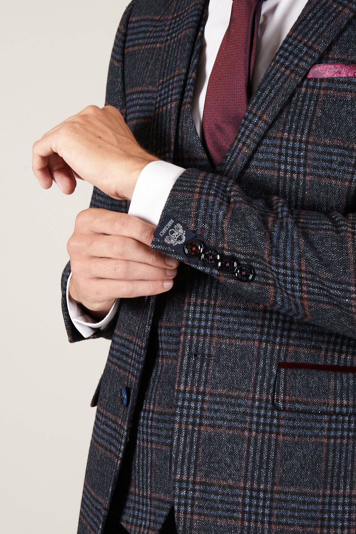 A man in a marcdarcy LUCA Navy Check Tweed Blazer, white shirt, and burgundy tie adjusts his cuff. The blazer’s grey-blue check with thin red lines pairs with a matching vest and pink pocket square—perfect for vintage enthusiasts.