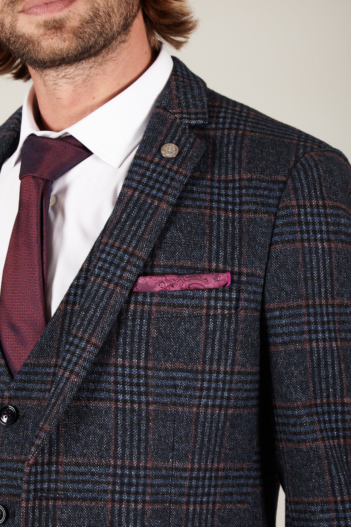 A man wears the marcdarcy LUCA Navy Check Tweed Blazer with a burgundy patterned tie, white pin collar shirt, pink paisley pocket square, and a visible silver lapel pin.