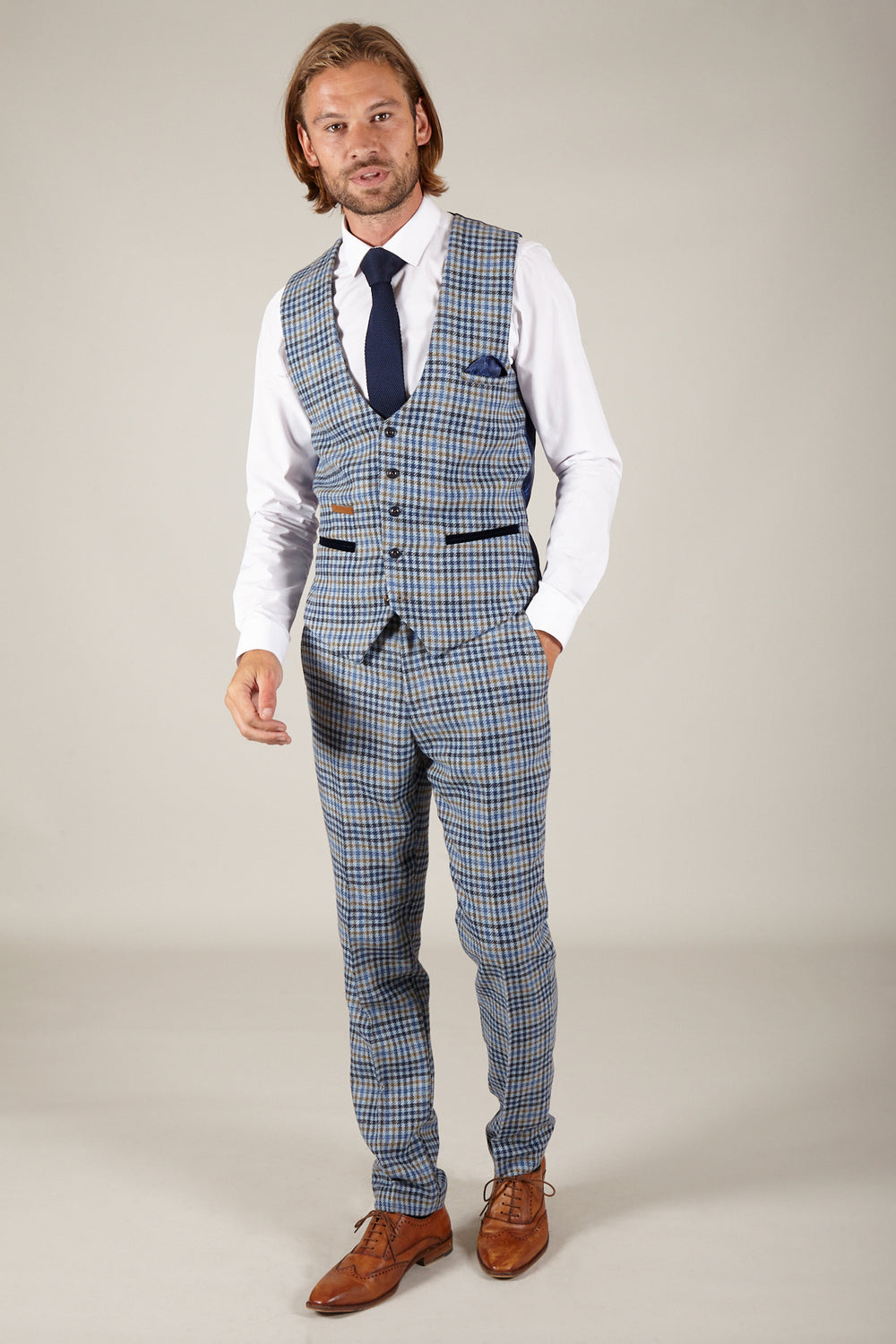 A man in a marcdarcy WATSON Blue Tan Tweed Check Single Breasted Waistcoat, white shirt, and dark blue tie poses with his hand in his pocket against a neutral background.