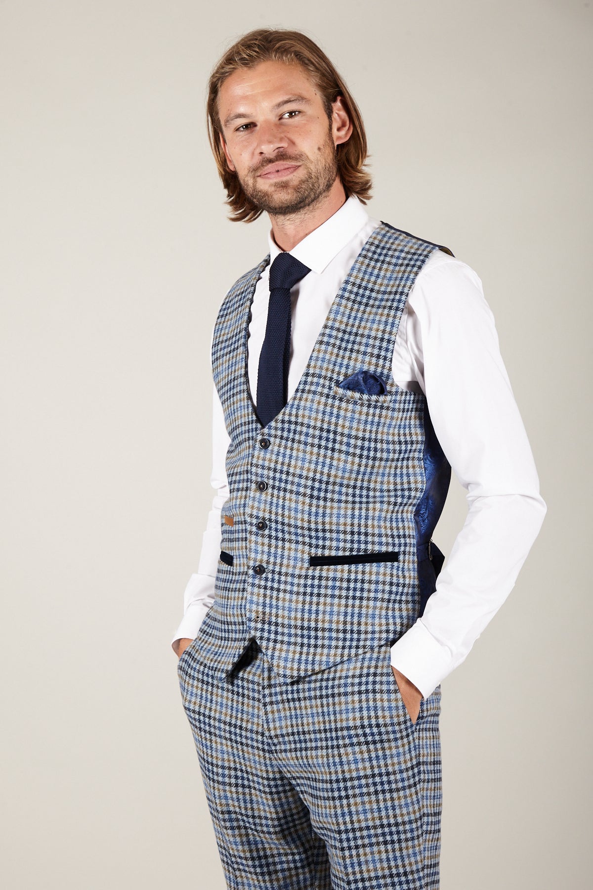 A man with shoulder-length hair and a beard wears the marcdarcy WATSON Blue Tan Tweed Check Single Breasted Waistcoat, matching trousers, a white shirt, navy tie, and pocket square, standing hands in pockets against a plain background.
