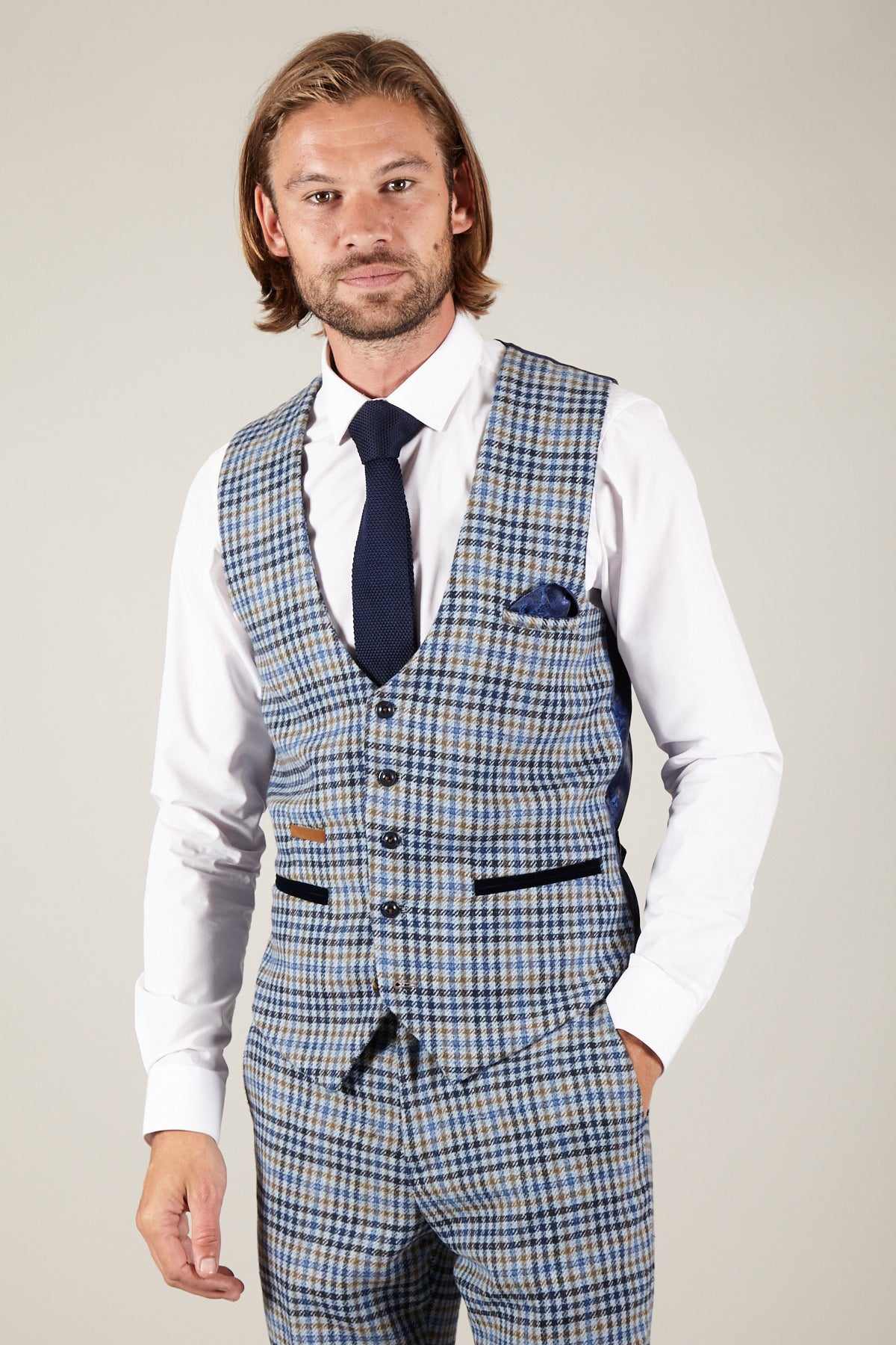 A man with shoulder-length hair and a beard wears the marcdarcy WATSON Blue Tan Tweed Check Single Breasted Waistcoat, matching pants, a white shirt, navy tie, and pocket square. He stands facing the camera against a neutral background.