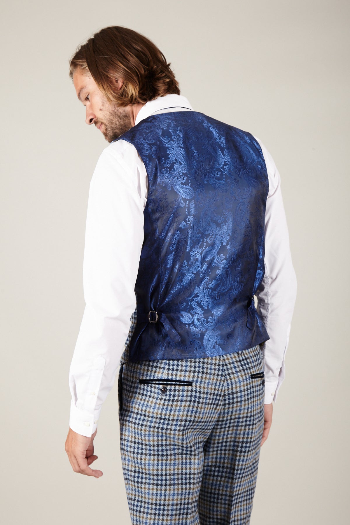 A man in a slim fit white shirt and blue plaid trousers stands with his back to the camera, highlighting the marcdarcy WATSON Blue Tan Tweed Check Single Breasted Waistcoat with side adjusters and paisley print lining as he looks over his shoulder.