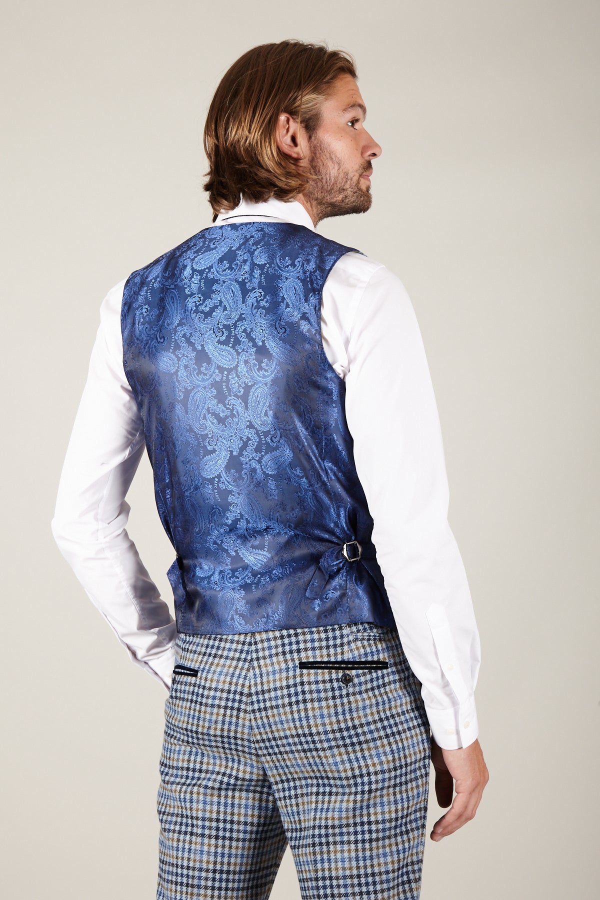 A man with shoulder-length light brown hair faces away, wearing a white shirt, gray plaid pants, and the marcdarcy WATSON Blue Tan Tweed Check Single Breasted Waistcoat featuring an adjustable side buckle and slim fit for a British-inspired style.