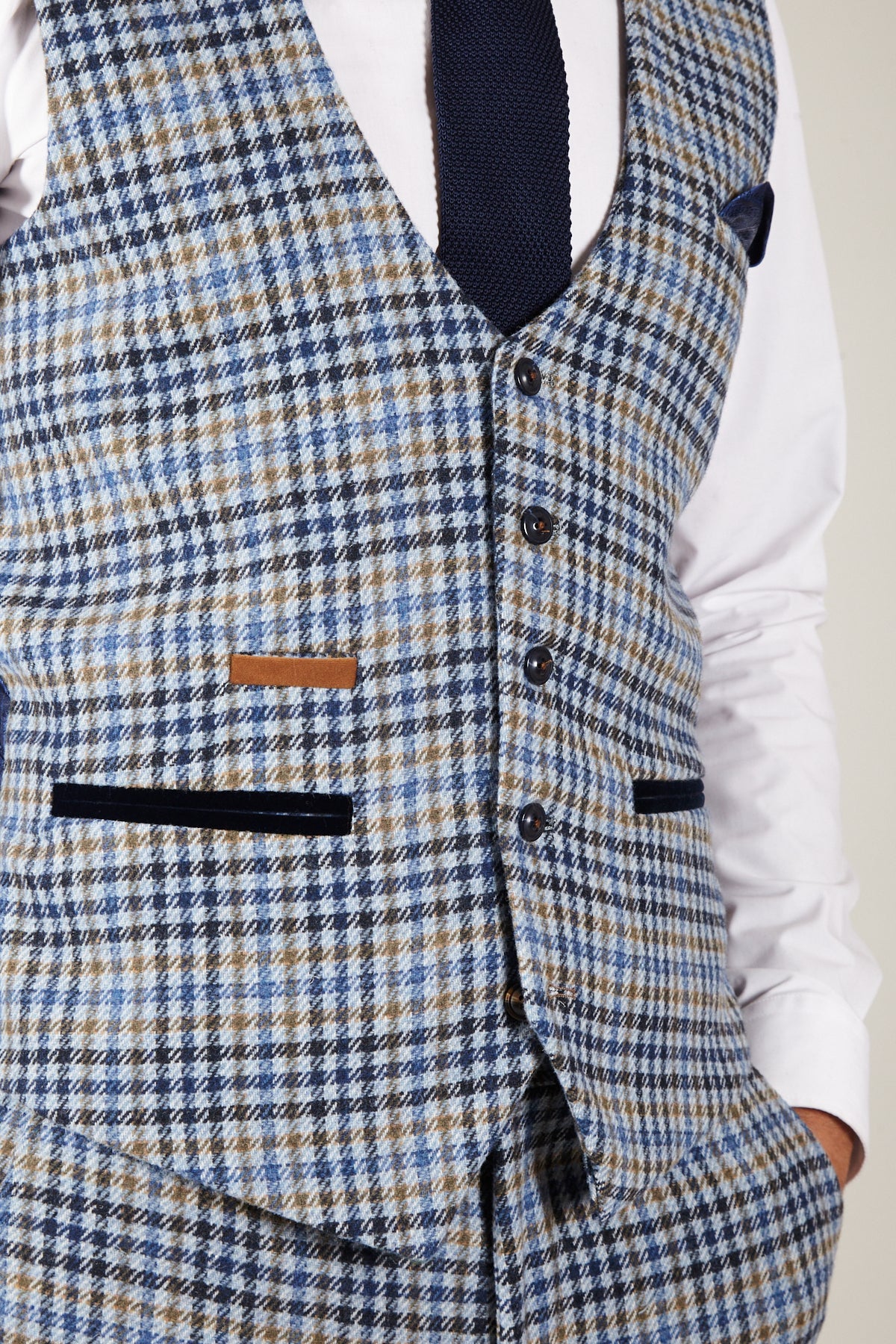 A man wears the marcdarcy WATSON Blue Tan Tweed Check Single Breasted Waistcoat with matching trousers, white shirt, and navy tie. The vest has black buttons, pocket details, a brown accent, and paisley lining.