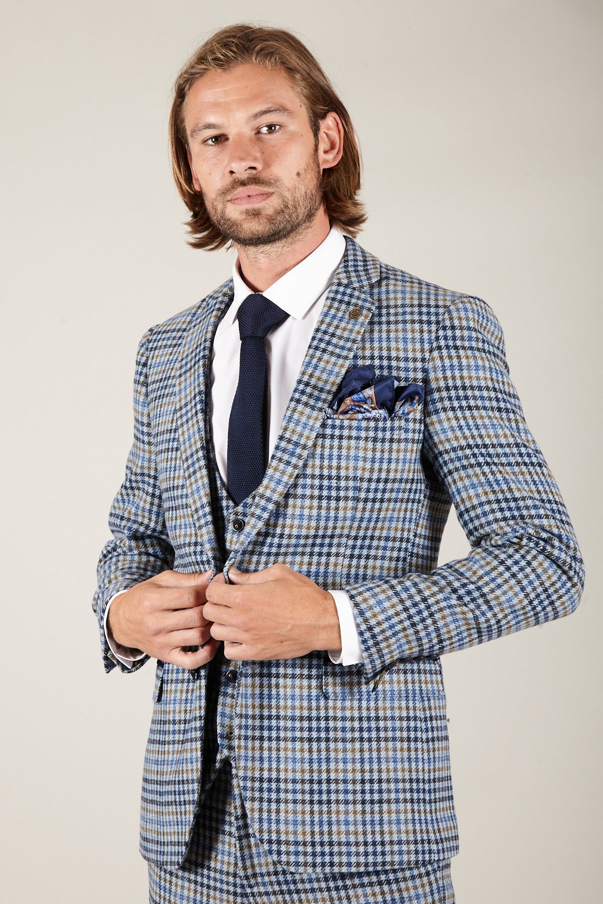 A man with shoulder-length hair wears the Marc Darcy WATSON Blue Tan Tweed Check Blazer over a white shirt and dark tie, standing against a plain background while buttoning his jacket.