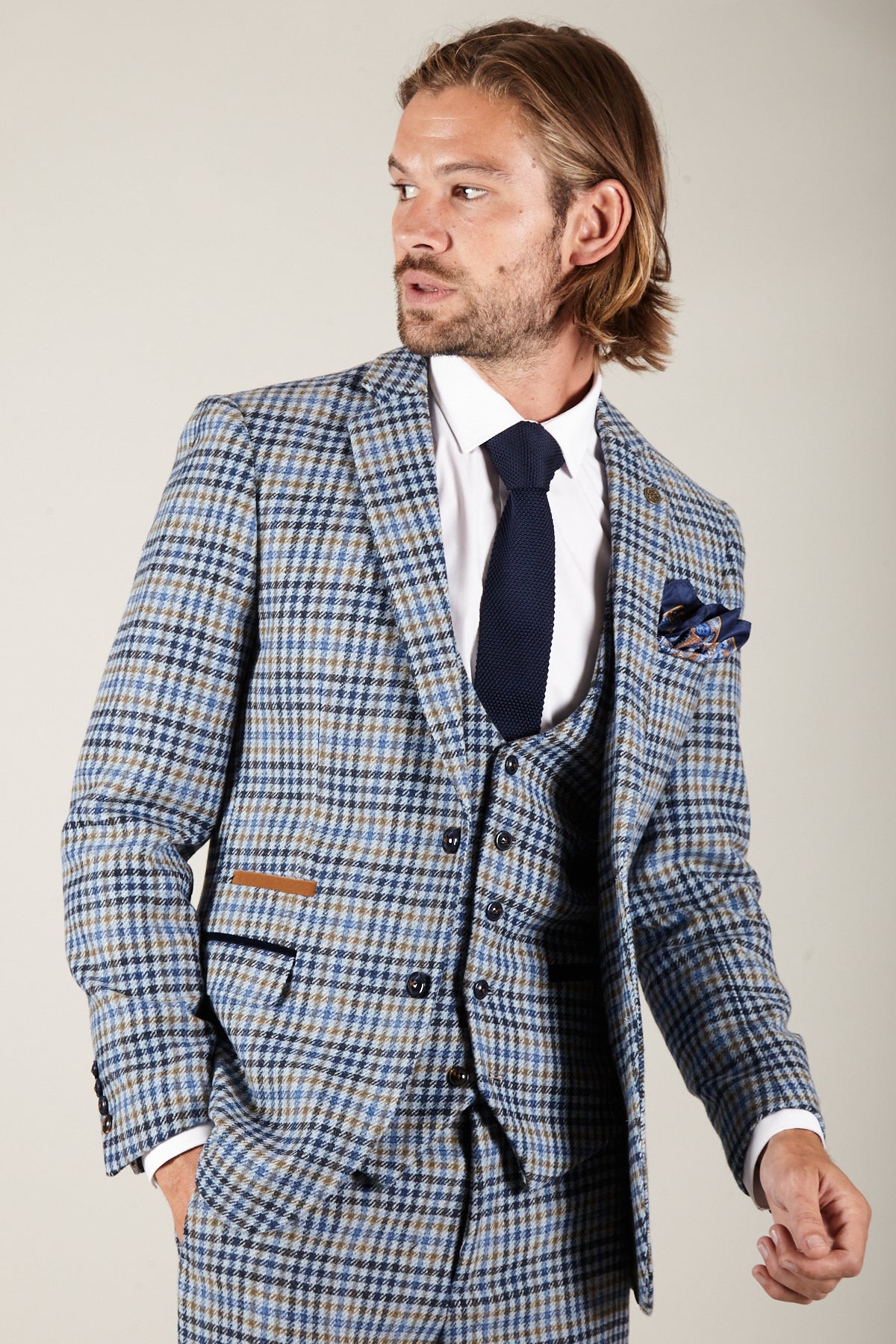 A man with shoulder-length hair and a beard wears the Marc Darcy WATSON Blue Tan Tweed Check Blazer over a white shirt and dark tie, looking left. A patterned pocket square hints at the paisley lining inside.