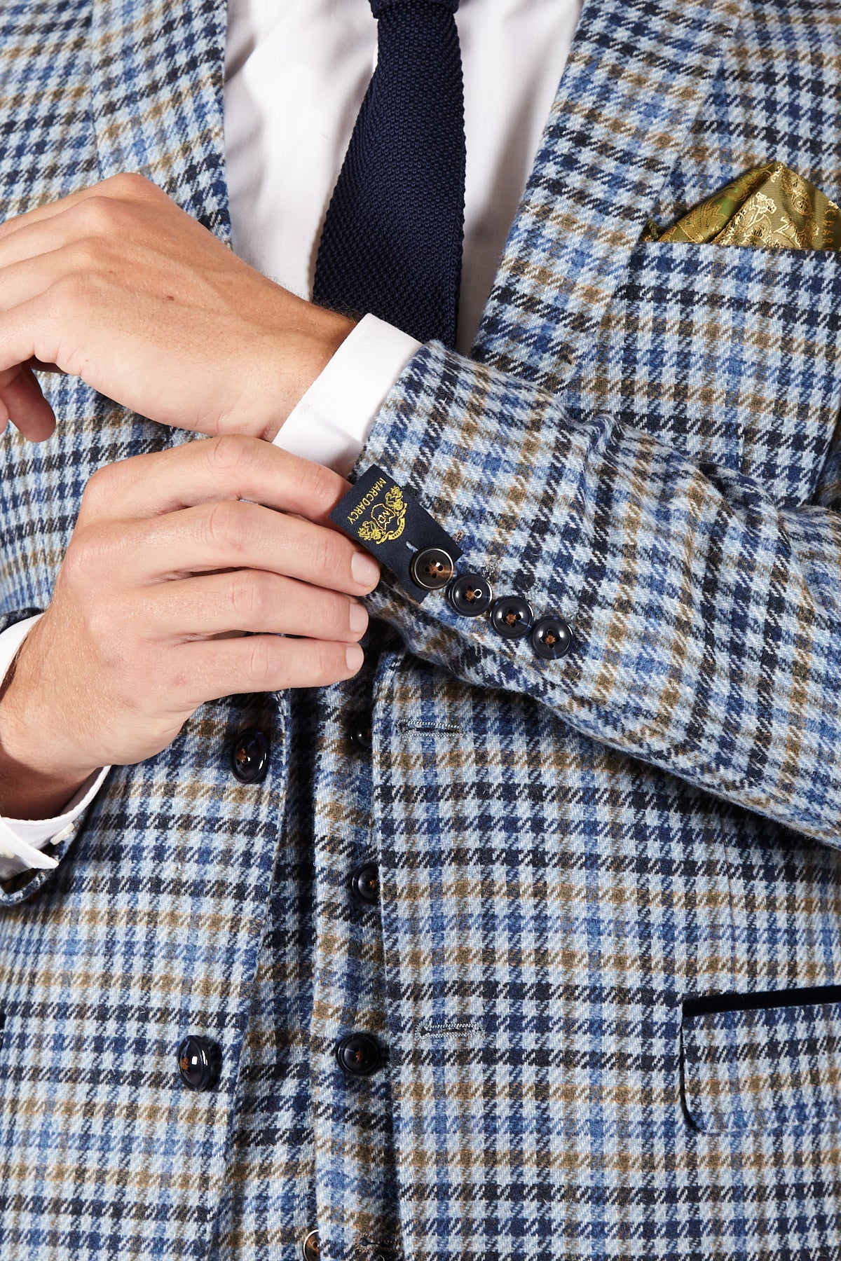 A person adjusts their sleeve, revealing a label on a Marc Darcy WATSON Blue Tan Tweed Check Blazer, styled with a white shirt, navy tie, and yellow pocket square. Only their upper body and hands are visible.