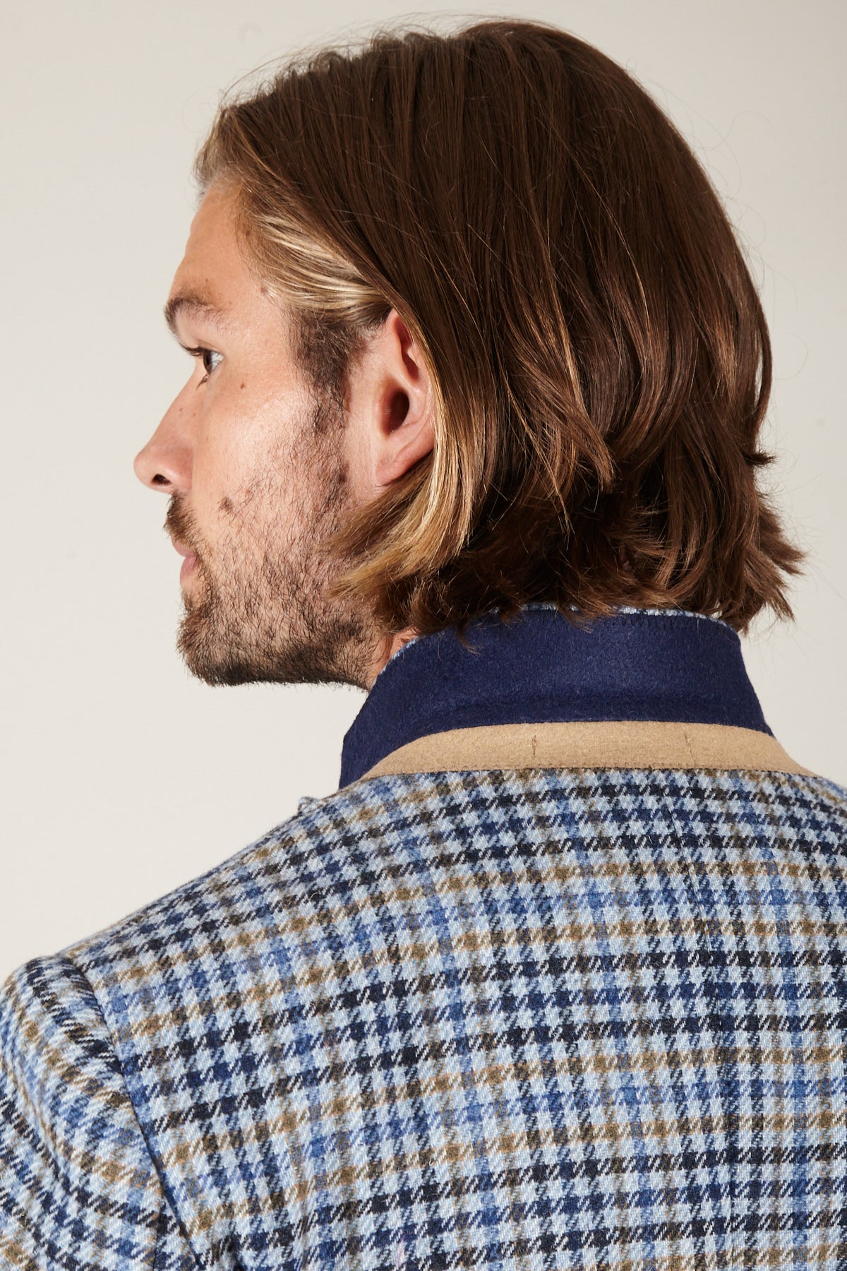 A man's profile, looking left, shows his long brown hair, beard, and a Marc Darcy WATSON Blue Tan Tweed Check Blazer with a navy collar—capturing classic British style.