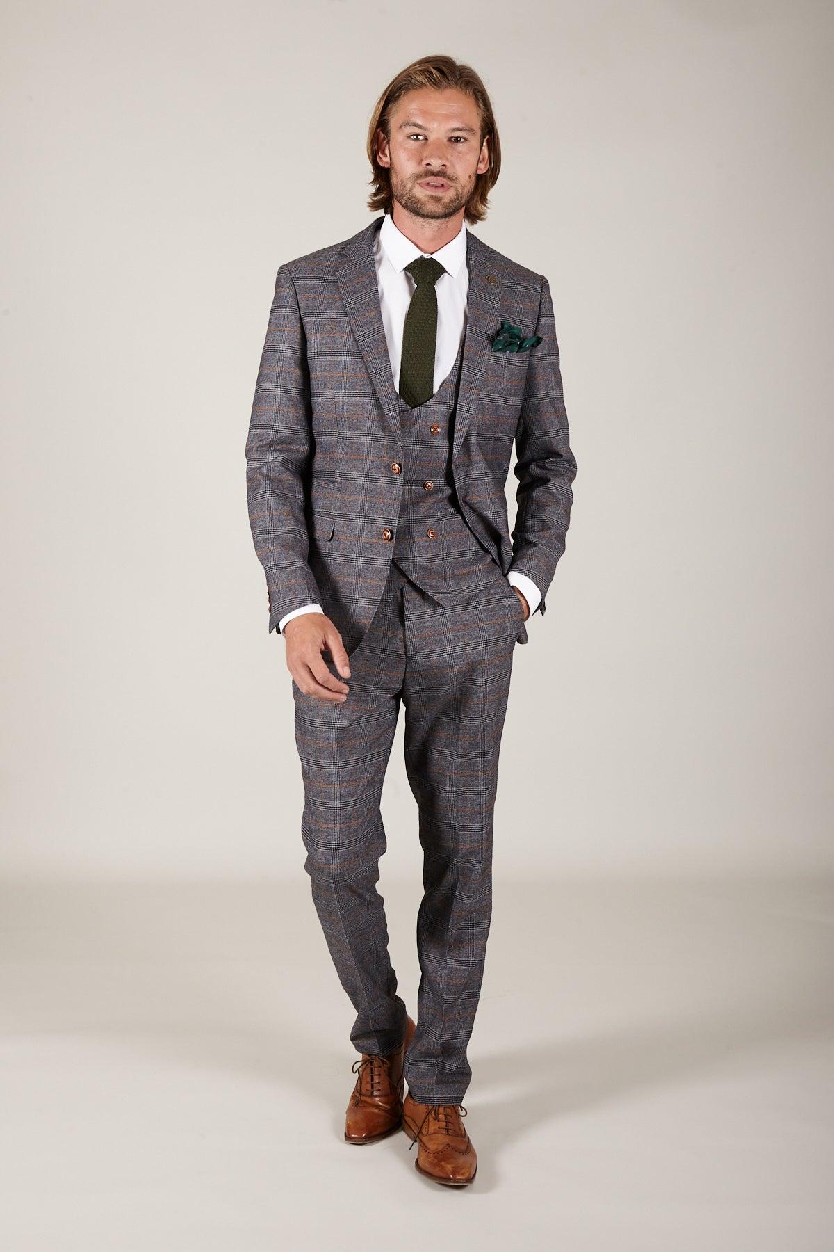 Dominic Calvert-Lewin wears The JENSON Grey Suit from marcdarcys Everton Collection—a slim fit three-piece suit with white shirt and dark tie, styled with shoulder-length hair, a beard, and brown dress shoes against a light background.