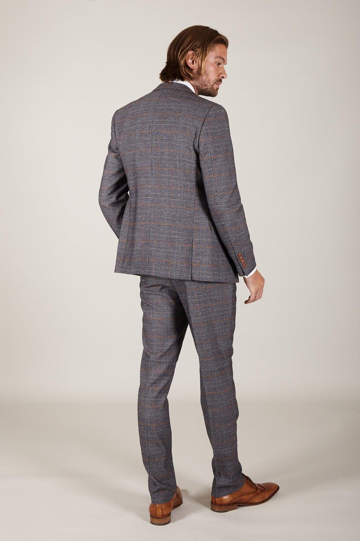 A man with shoulder-length hair faces away, wearing the marcdarcy JENSON Grey Check Suit with KELVIN Tan Waistcoat and brown dress shoes, glancing over his shoulder against a plain light background.