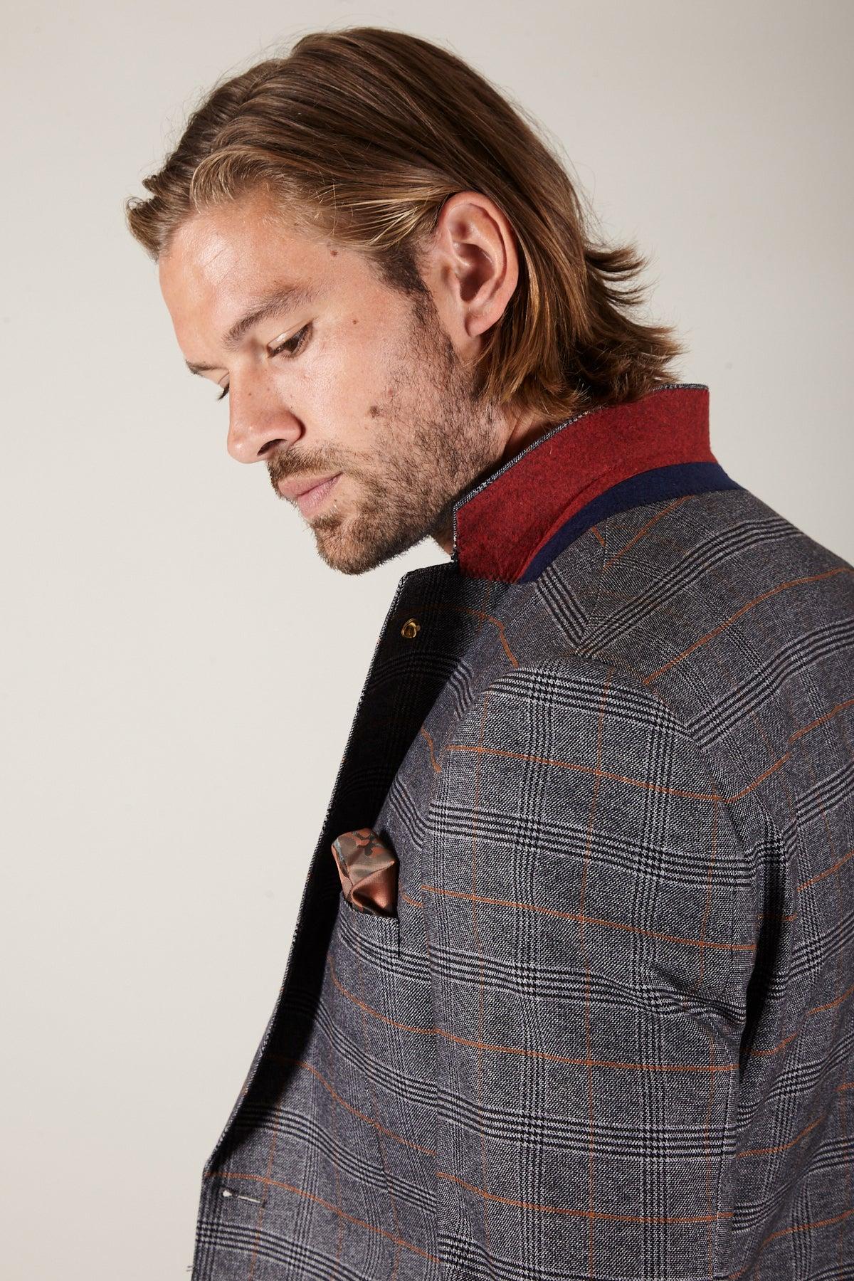 A man with shoulder-length hair and a beard wears the marcdarcy JENSON grey check suit with KELVIN tan waistcoat, styled with a red collar and pocket square, looking down against a plain background.