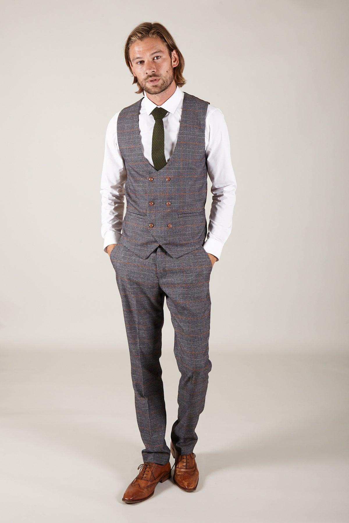 A man with shoulder-length hair models marcdarcy’s The Everton Collection | Dominic Calvert-Lewin in the JENSON Grey Suit, pairing it with a white shirt, green tie, and brown shoes, standing hands-in-pockets against a light background.