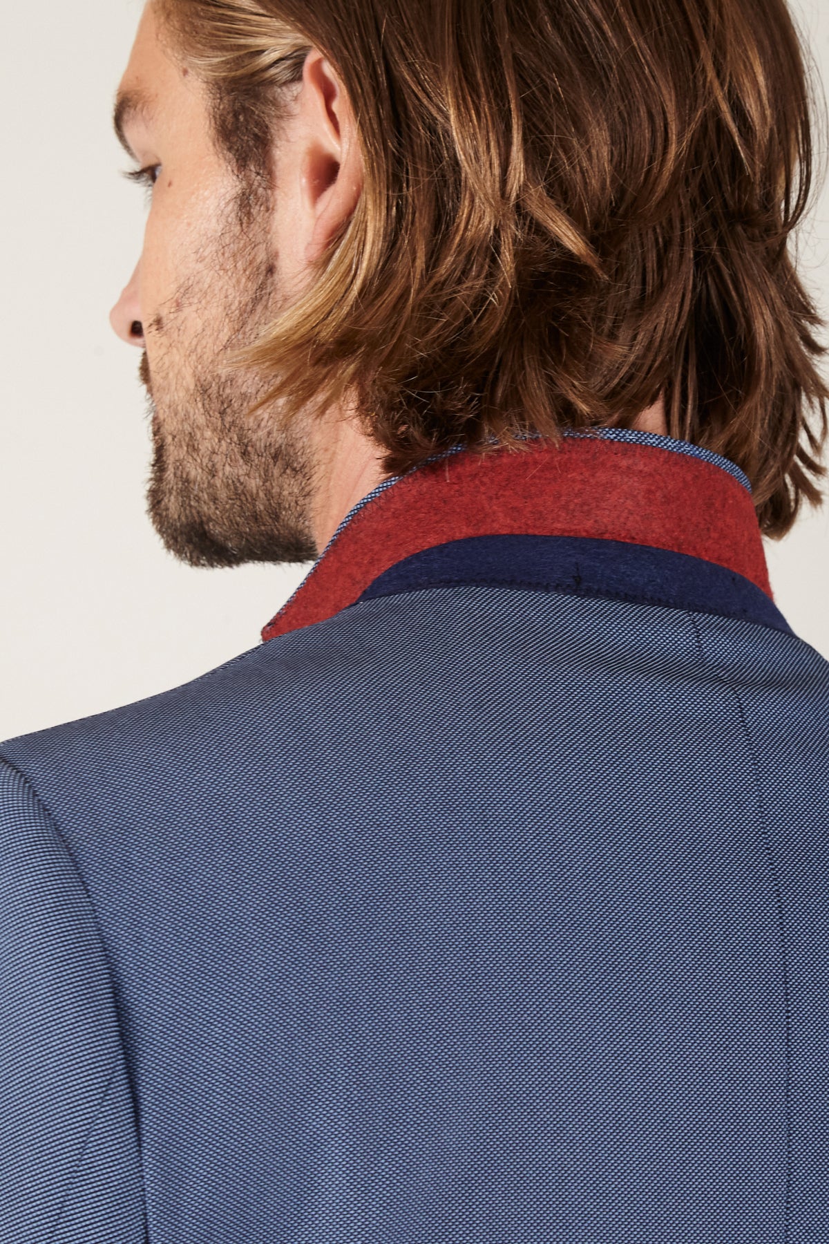 A man with light brown, shoulder-length hair and a beard is seen from behind wearing the Marc Darcy DANNY - Sky Tailored Blazer, featuring blue textured fabric, red and blue collar accents, slim fit, and functional pockets.