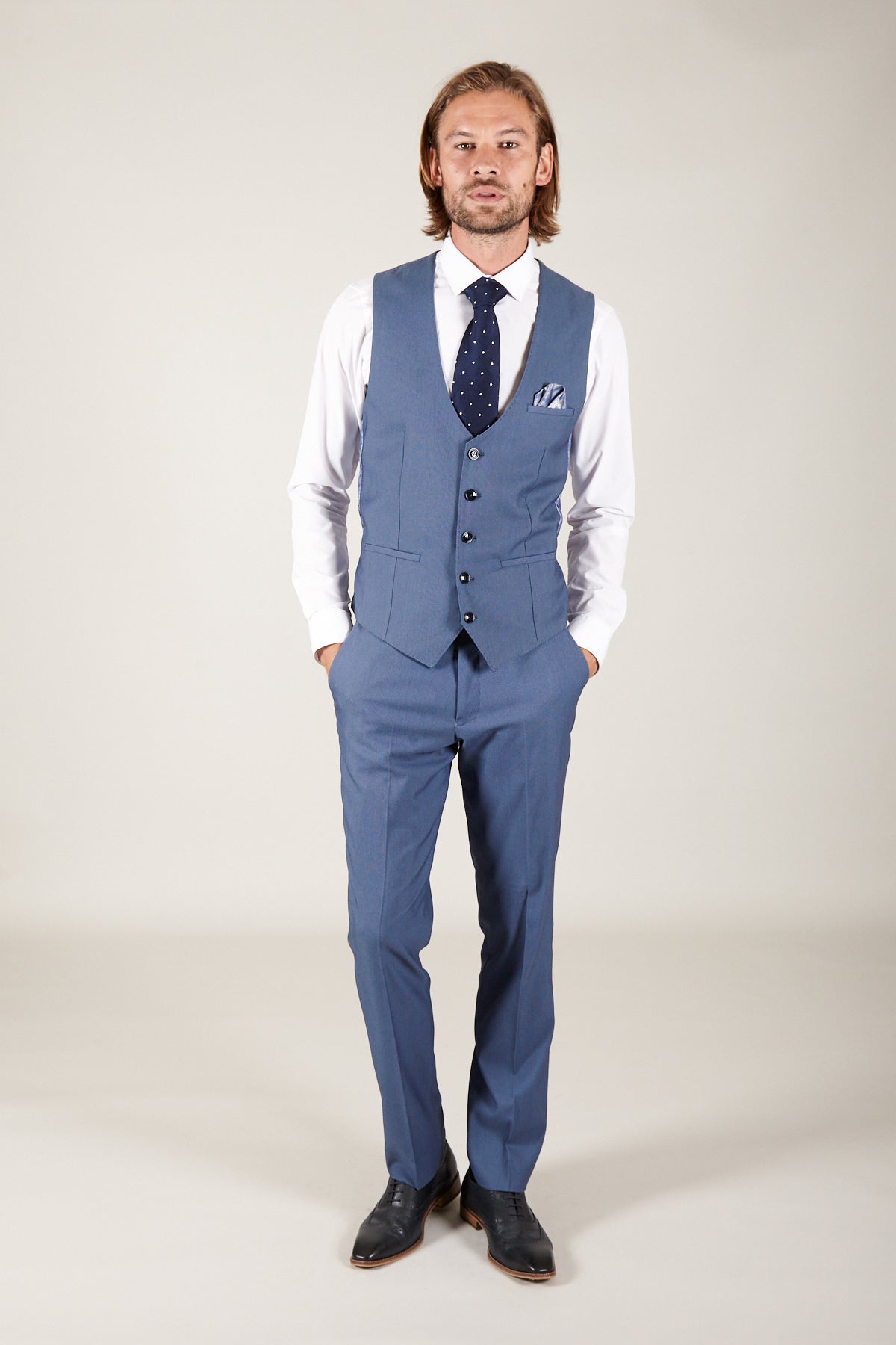A man with light brown hair and a beard wears the Marc Darcy DANNY - Sky Single Breasted Waistcoat, paired with a white shirt, polka-dot tie, and black shoes. He stands hands-in-pockets against a plain background.
