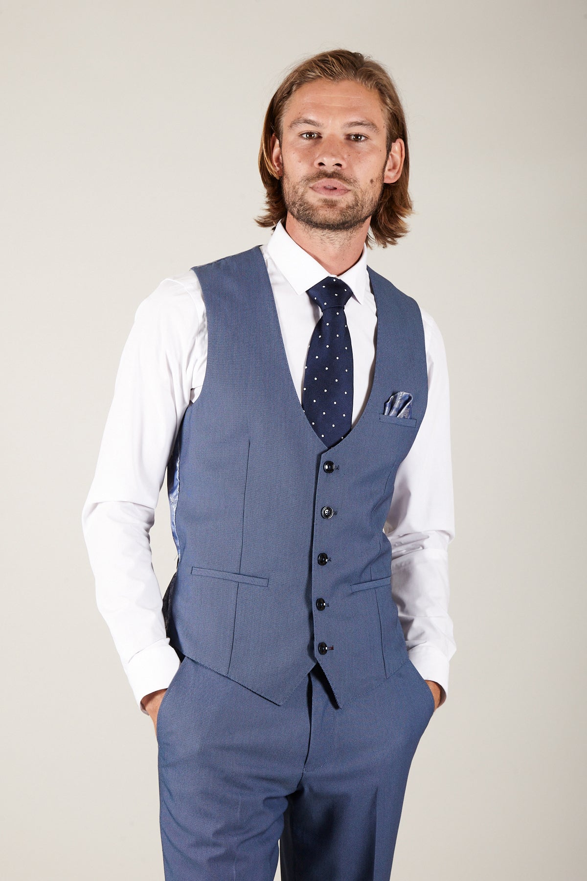 A man with shoulder-length light brown hair and a beard models the Marc Darcy DANNY - Sky Single Breasted Waistcoat, styled without the jacket over a white shirt, navy polka dot tie, and matching pocket square, hands in his trouser pockets.