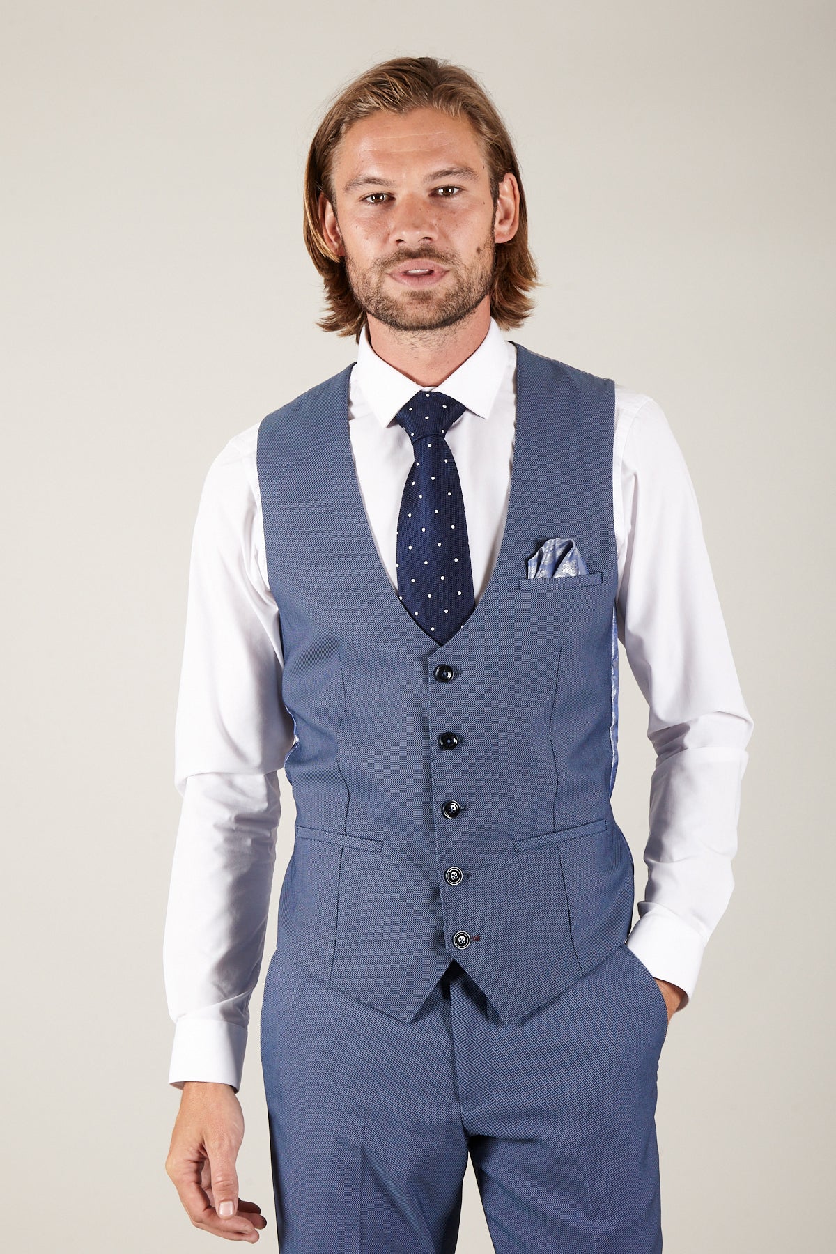 A man with long, light brown hair and a beard wears the Marc Darcy DANNY Sky Single Breasted Waistcoat and matching trousers, a white shirt, and a navy polka-dot tie, standing against a plain light background with one hand in his pocket.