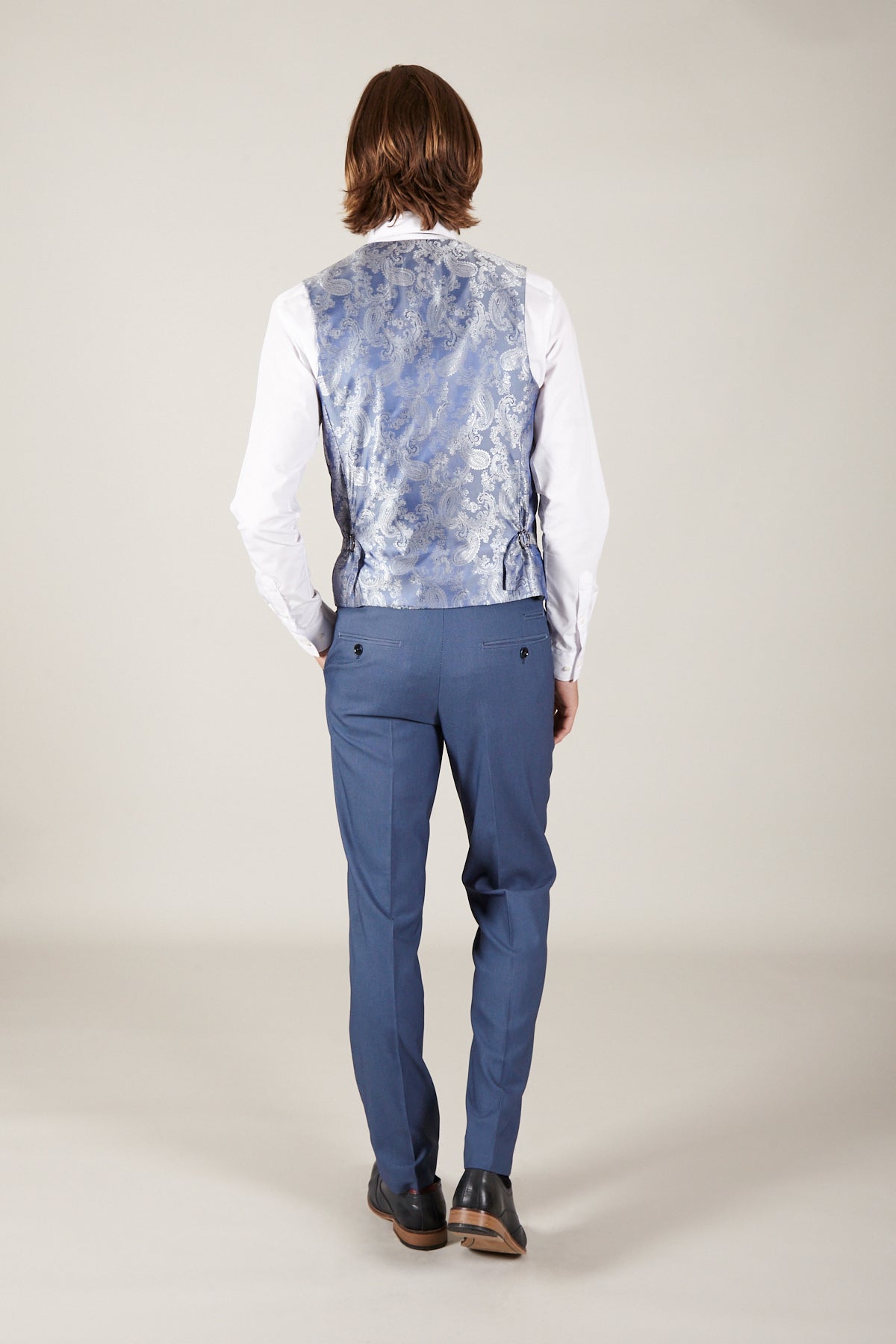 A person with medium-length brown hair, dressed in the Marc Darcy DANNY - Sky Single Breasted Waistcoat, white shirt, blue dress pants, and brown shoes stands facing away from the camera against a plain light background.