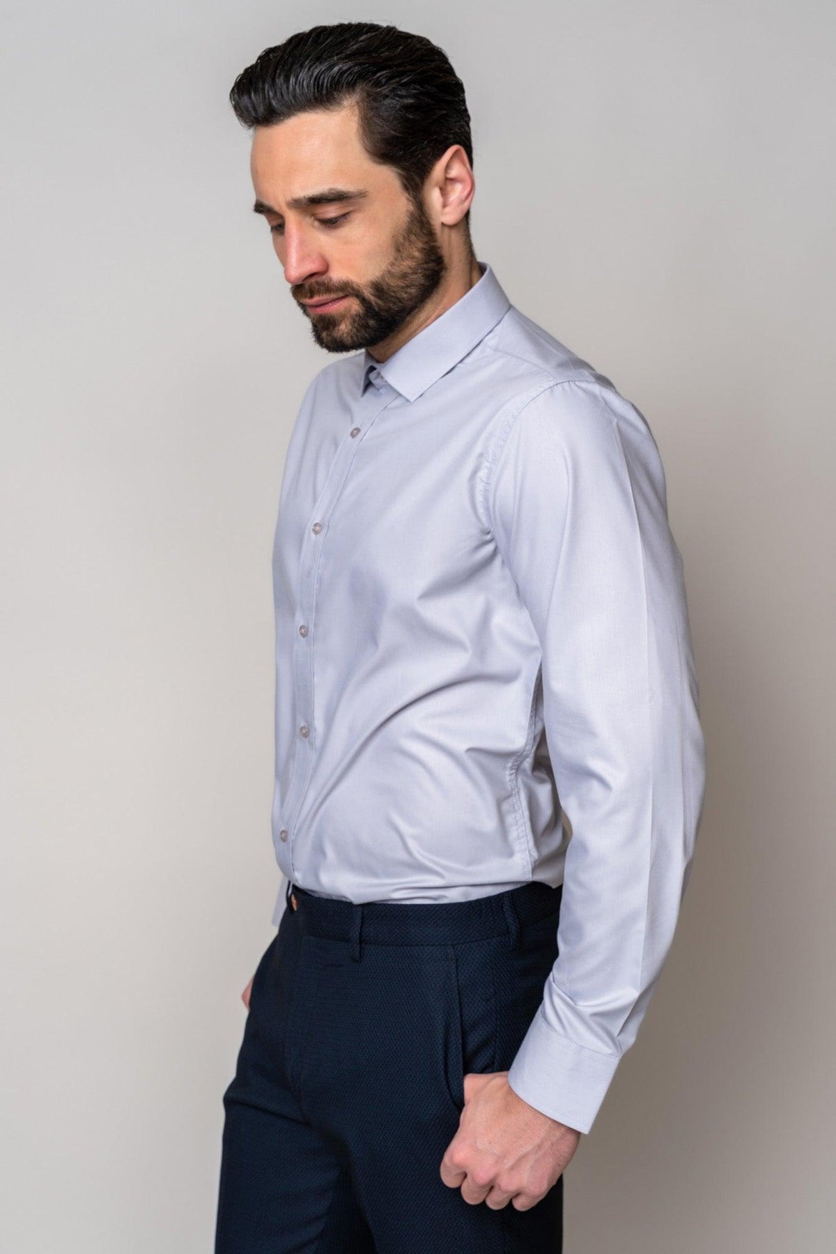 A man with dark hair and a beard, wearing the CARTER - Silver Long Sleeve Shirt by Marc Darcy and dark patterned pants, stands looking down.
