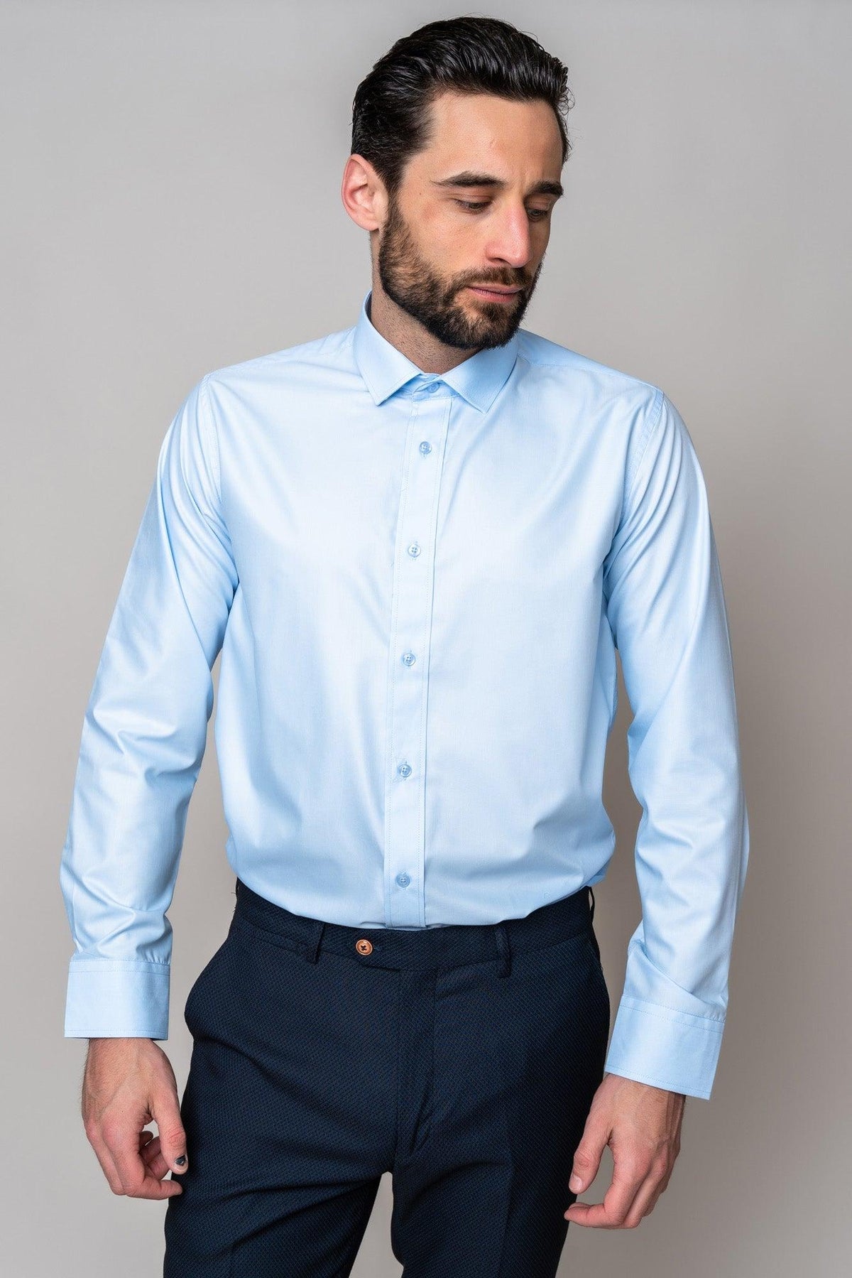 A man with dark hair and a beard wears the Marc Darcy CARTER - Sky Long Sleeve Shirt paired with dark trousers, standing against a plain gray background and looking slightly down to his left.