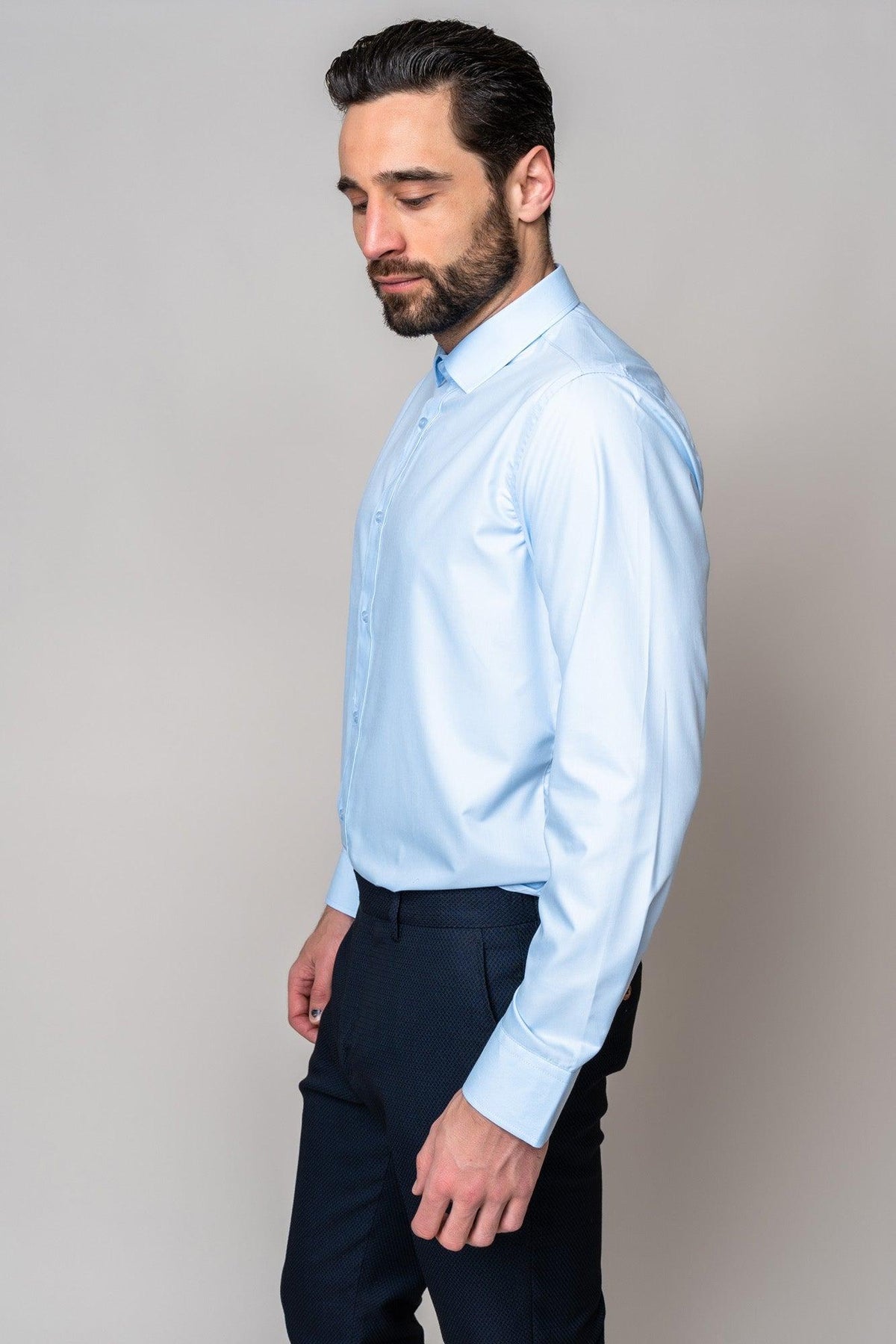 A man with dark hair and a beard stands sideways, wearing the Marc Darcy CARTER - Sky Long Sleeve Shirt with a spread collar and dark trousers, against a plain light gray background.