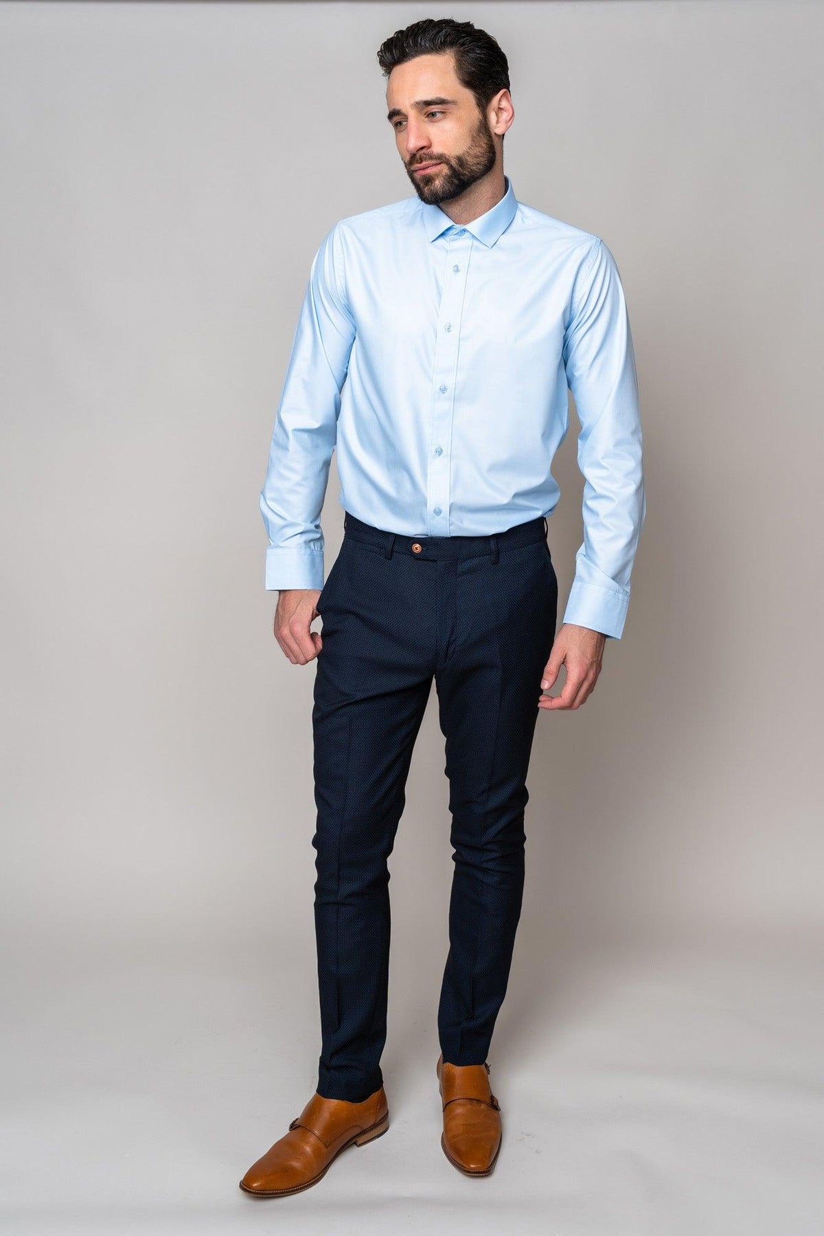 A man with dark hair and a beard stands against a plain background, wearing the Marc Darcy CARTER - Sky Long Sleeve Shirt, paired with dark navy trousers and brown dress shoes. He gazes slightly to the side in a relaxed pose.