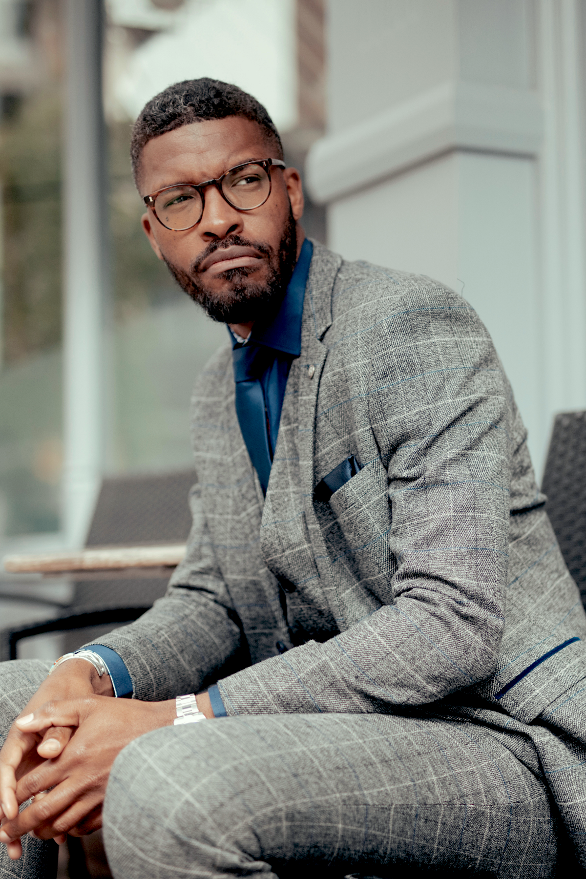 A Black man with a beard and glasses wears the marcdarcy SCOTT Grey Tweed Check Three Piece Suit, featuring heavyweight fabric and a slim fit, paired with a blue shirt. He looks to the side with a serious expression.