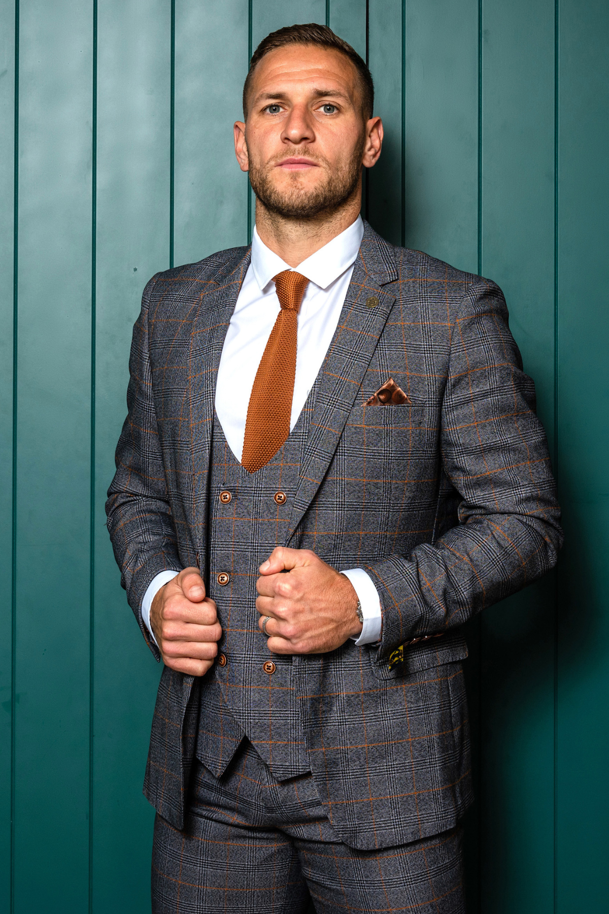 Billy Sharp models the marcdarcy Jenson Grey Check Suit from The Sheffield Utd Collection, standing confidently against a teal paneled wall, styled with a brown tie and pocket square, hands on his jacket.
