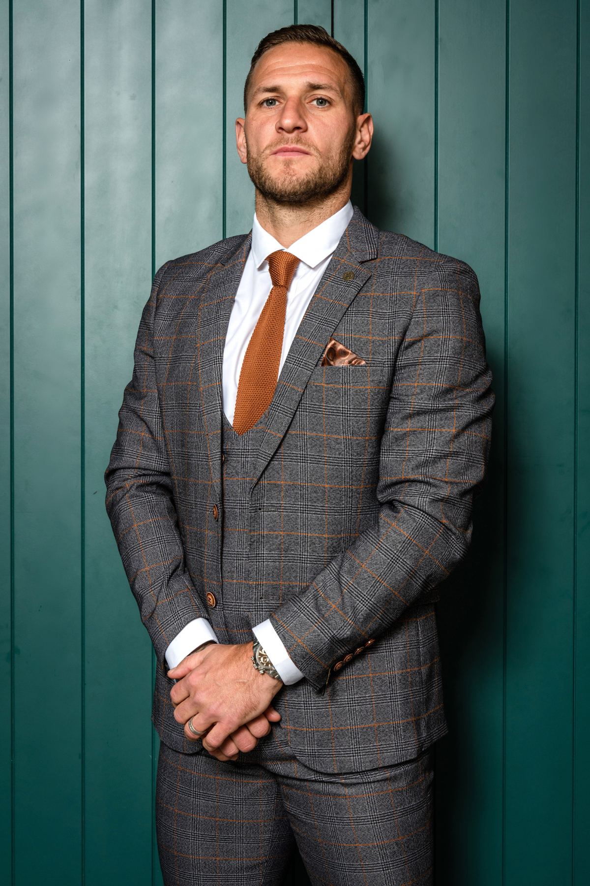 Billy Sharp wears the marcdarcy Jenson Grey Check Suit from The Sheffield Utd Collection, paired with a brown tie and pocket square, standing confidently before a green panel wall with his hands clasped.