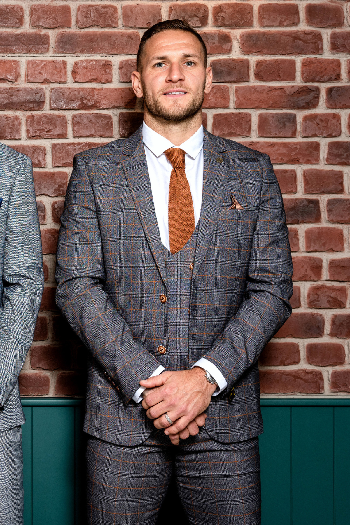 Billy Sharp wears the marcdarcy Jenson Grey Check Suit from The Sheffield Utd Collection, posing with clasped hands in front of a brick wall and looking at the camera.