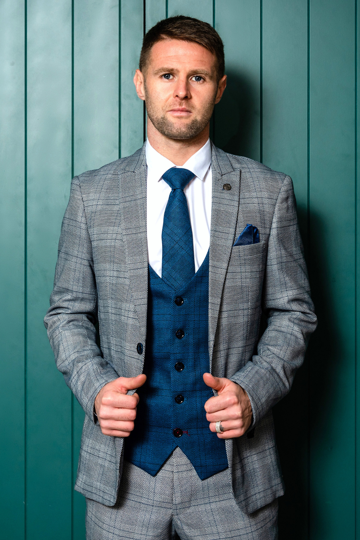 Oliver Norwood models the marcdarcy Jerry Check Suit from The Sheffield Utd Collection, pairing it with a white shirt and blue waistcoat against a teal backdrop, exuding sharp confidence as he looks into the camera.