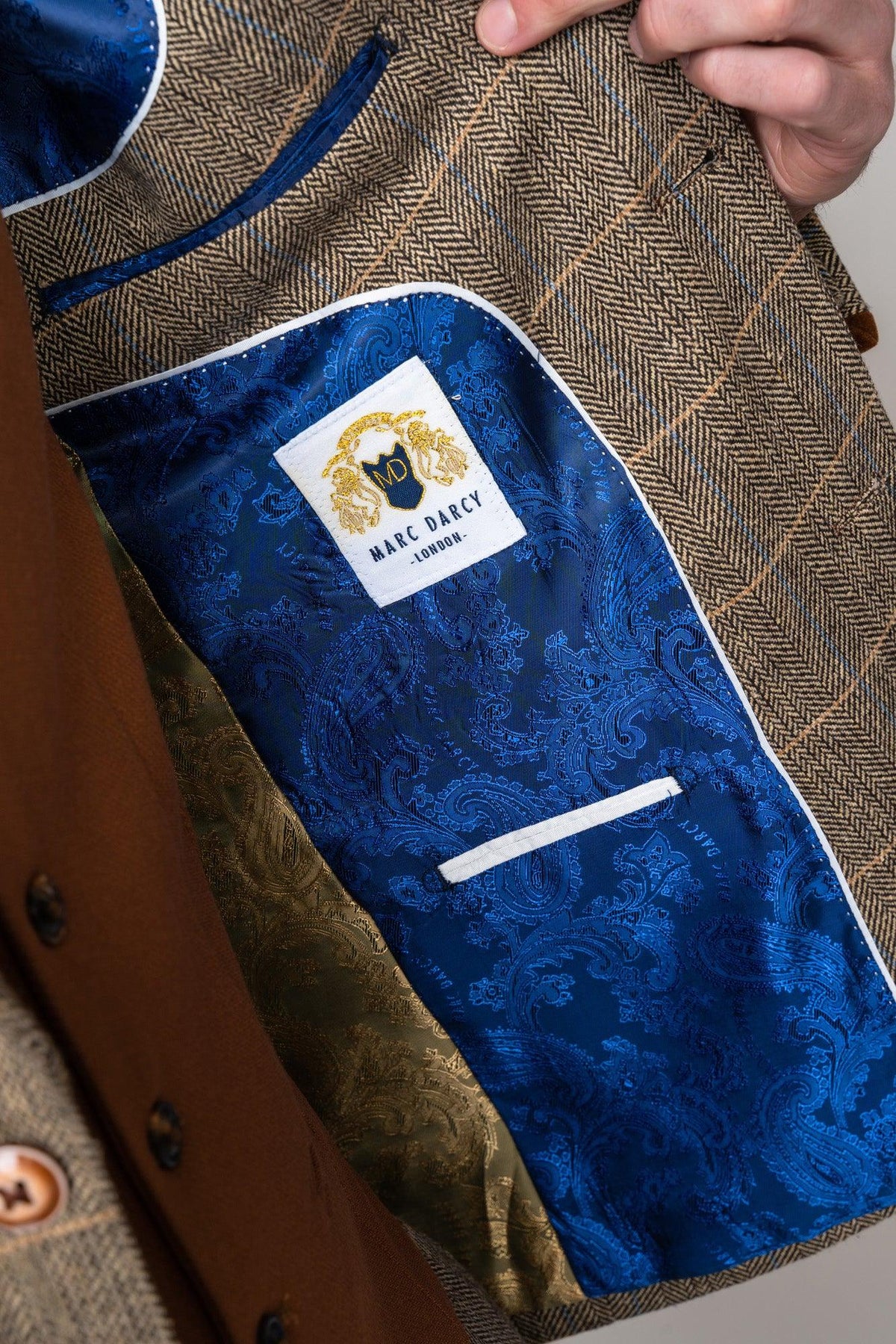 A close-up of someone holding open a marcdarcy TED - Tan Tweed Check Suit with Kelvin Tan Waistcoat, showing an inner pocket lined with shiny blue paisley fabric and a white brand label featuring a gold lion crest.
