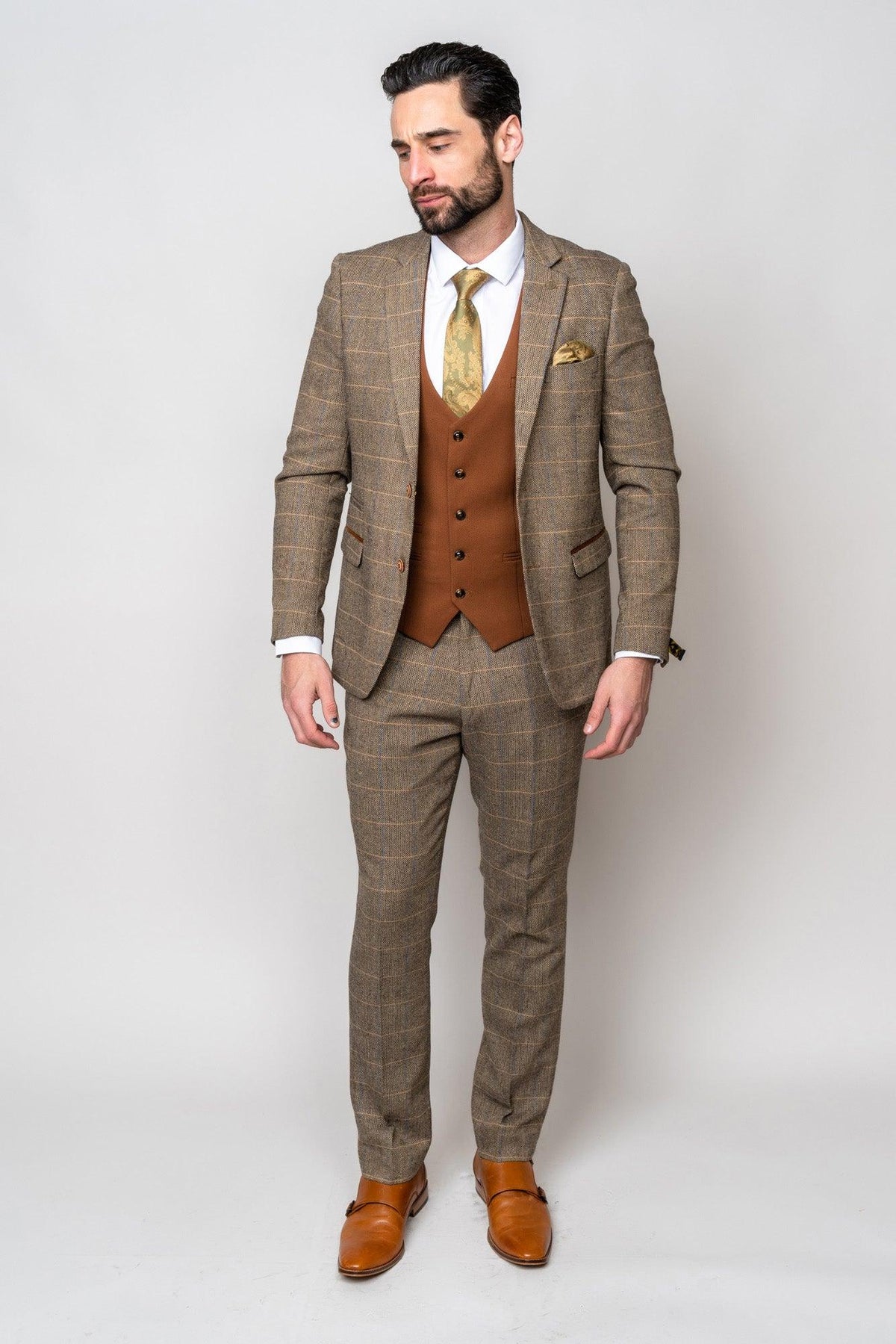 A man stands against a light background wearing the marcdarcy TED Tan Tweed Check Suit with matching Kelvin Tan Waistcoat, white shirt, patterned gold tie, and brown dress shoes, looking slightly downward with hands relaxed at his sides.