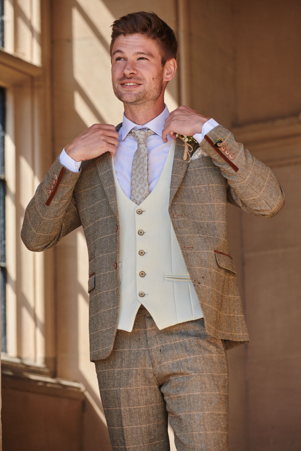 A man wearing the marcdarcy TED Tan Tweed Check Suit with a Kelvin Cream Waistcoat stands indoors, adjusting his jacket and smiling as sunlight streams in—ideal attire for country style weddings.