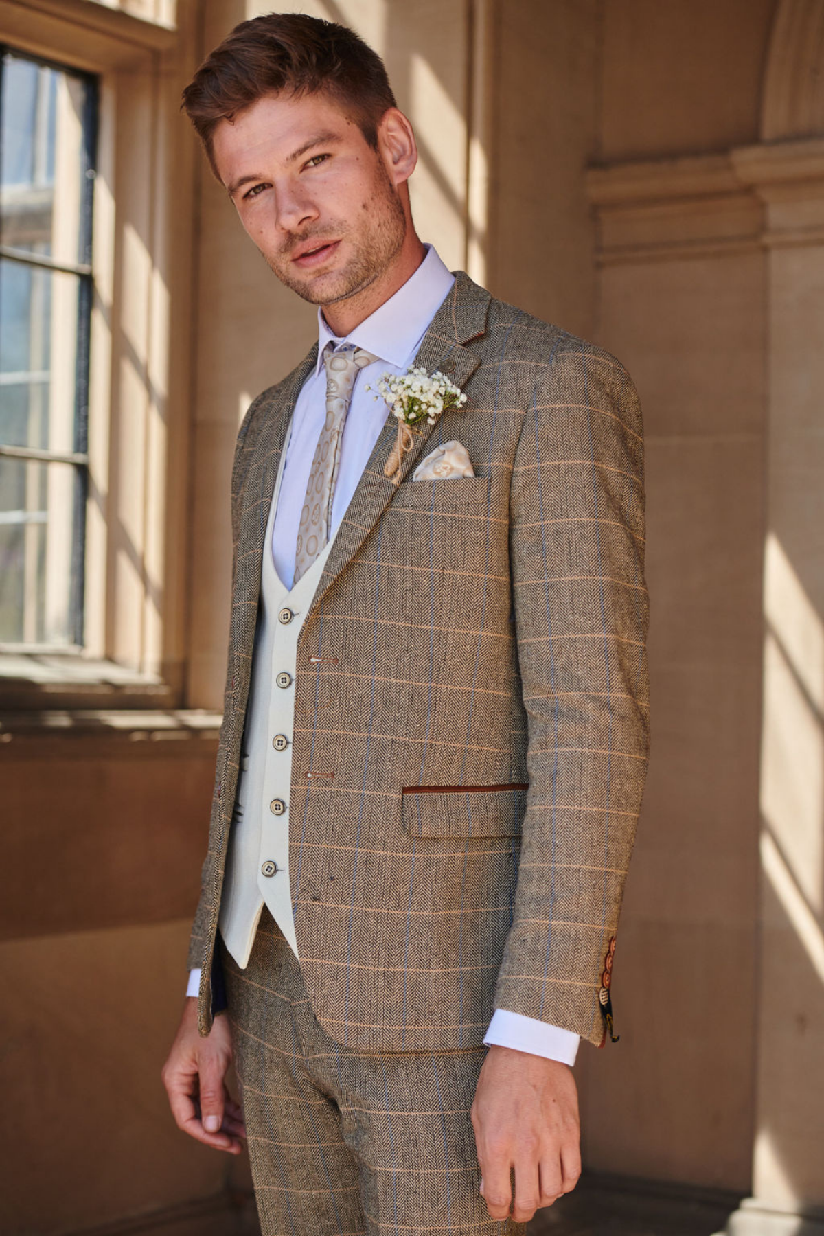 A man in the marcdarcy TED Tan Tweed Check Suit with Kelvin Cream Waistcoat stands by a window, styled with a white shirt, patterned tie, boutonniere, and pocket square—perfect for country-style weddings.