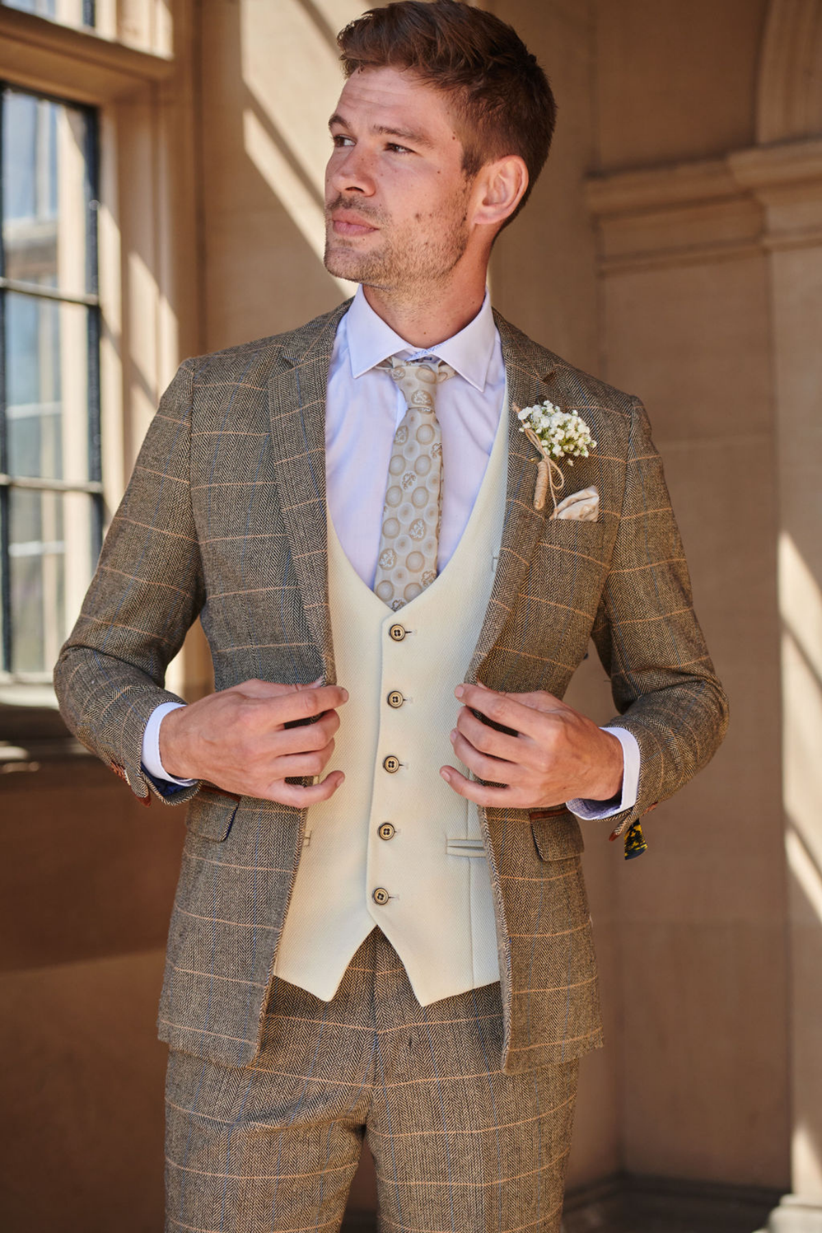 A man in the marcdarcy TED Tan Tweed Check Suit with Kelvin Cream Waistcoat adjusts his jacket near a window, a patterned tie and boutonnière completing his look—ideal for country style weddings.