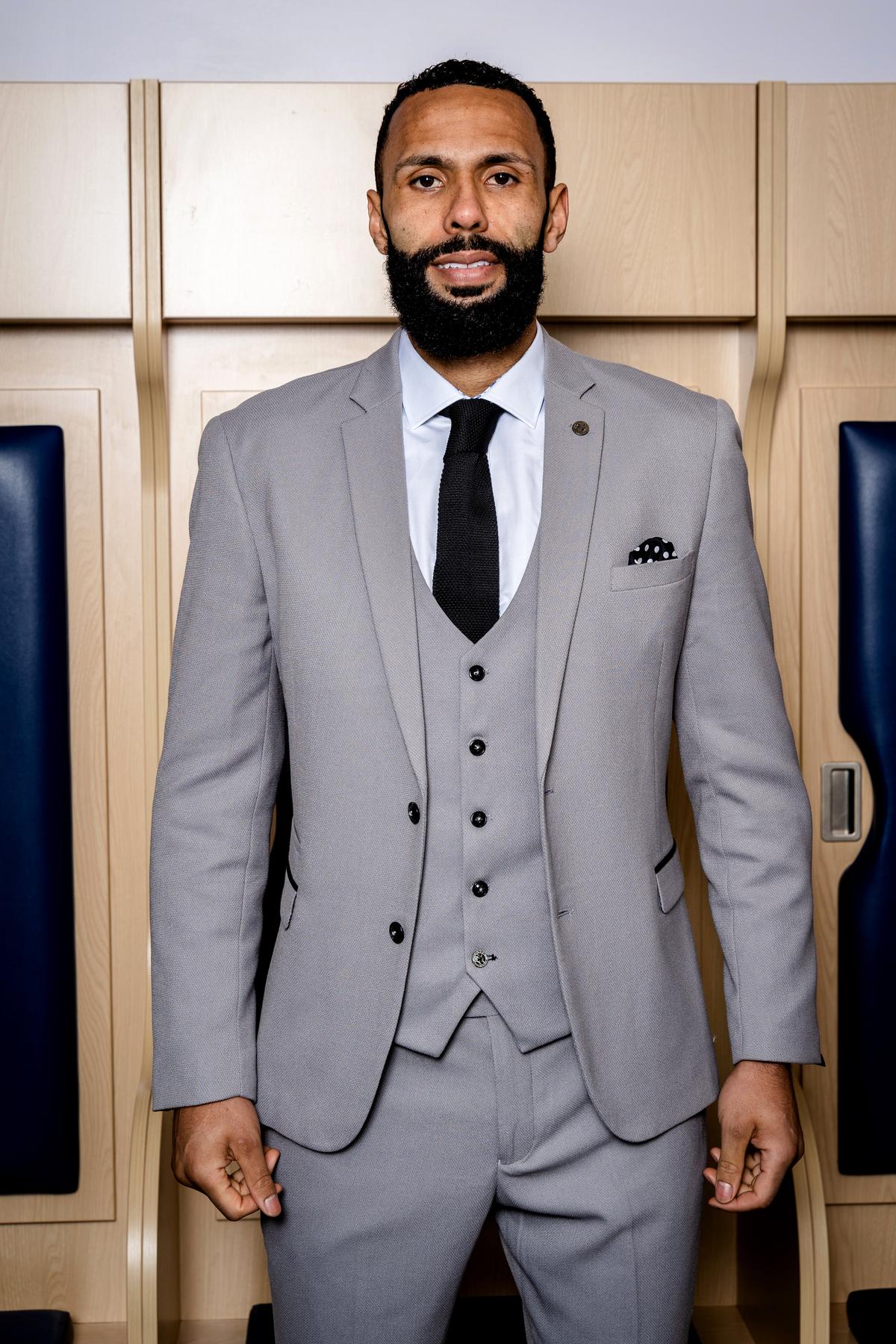A bearded man wears the marcdarcy WBA Collection - Edwin Suit as worn by Kyle Bartley in light gray with a white shirt, black tie, and pocket square, standing before wooden lockers and looking at the camera.