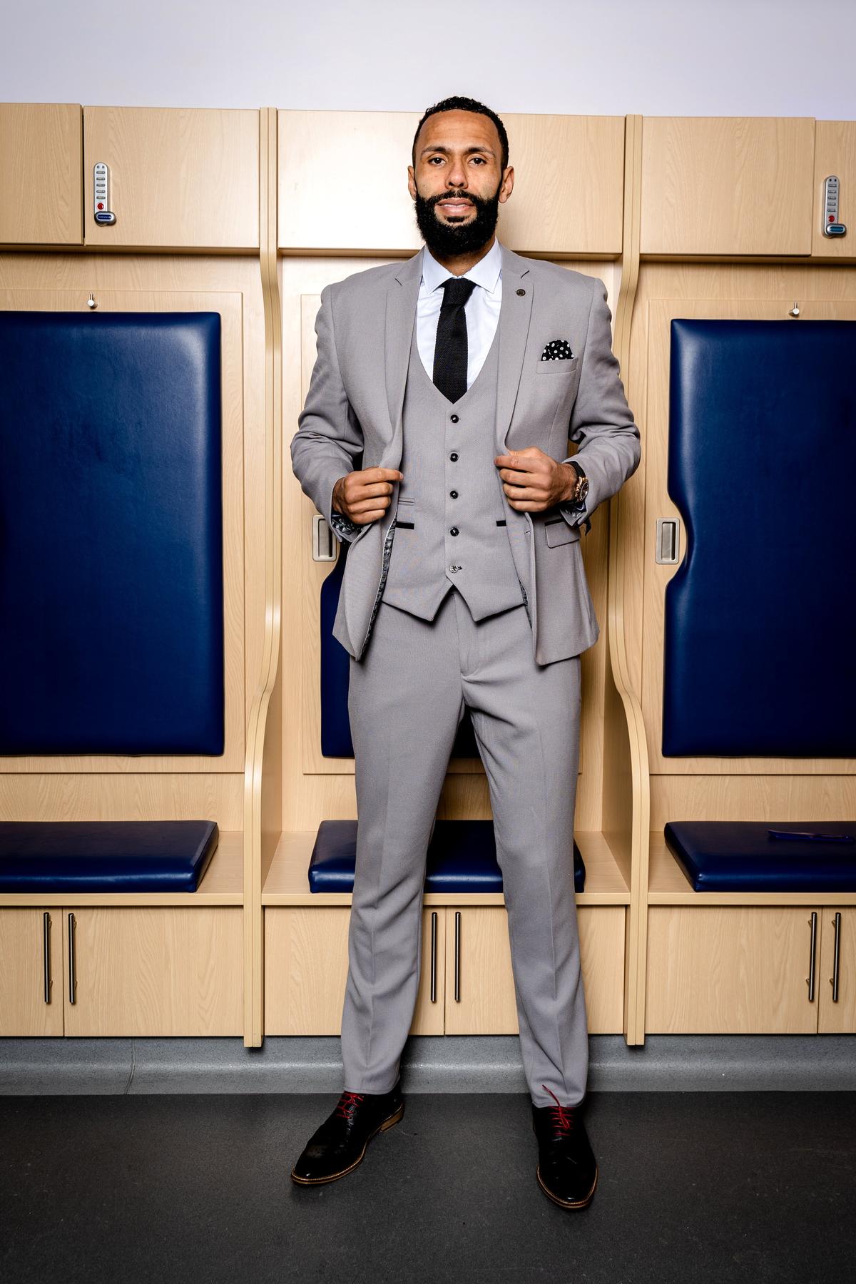 A man in the marcdarcy WBA Collection Edwin Suit as worn by Kyle Bartley, light grey and slim fit with a black tie, stands in a locker room holding open his jacket.