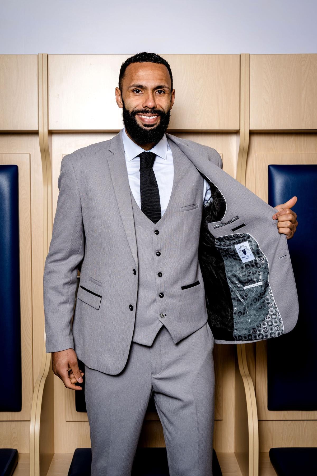 A man in The WBA Collection - Edwin Suit by marcdarcy, as worn by Kyle Bartley, smiles in a slim fit light grey jacket with a white shirt and black tie, revealing its patterned lining.