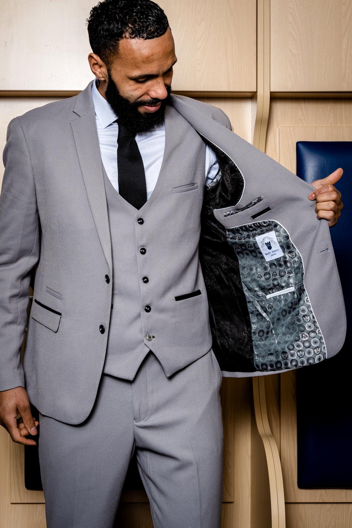 A man wears the marcdarcy Edwin Suit from The WBA Collection, as worn by Kyle Bartley—a slim fit light grey suit with a white shirt and black tie—smiling before wooden panels and revealing the jacket's patterned inner lining.