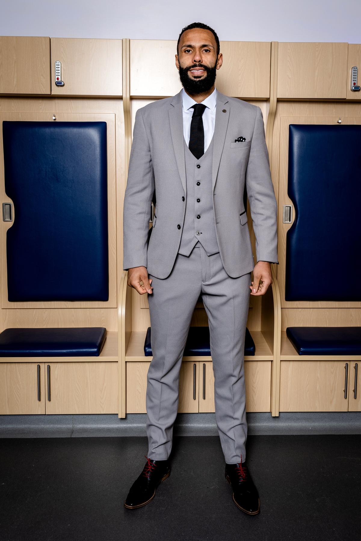 A man with a short beard and mustache smiles in front of wooden locker room cubicles, wearing marcdarcy’s The WBA Collection - Edwin Suit as worn by Kyle Bartley. He's styled with a black tie and black shoes featuring red laces.