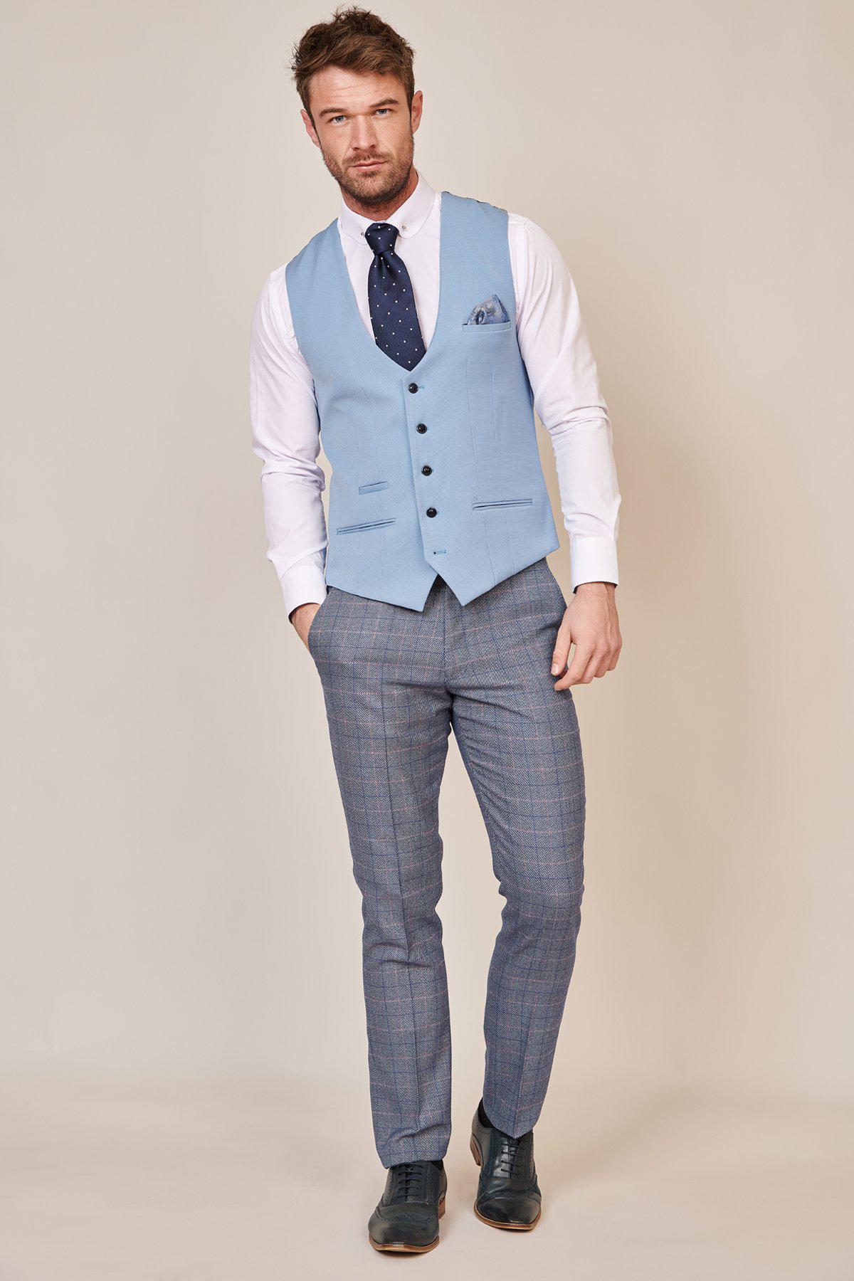 A man stands against a beige background wearing the marcdarcy KELLY Sky Blue Single Breasted Waistcoat over a white shirt, a navy polka-dot tie, gray plaid trousers, and dark shoes.