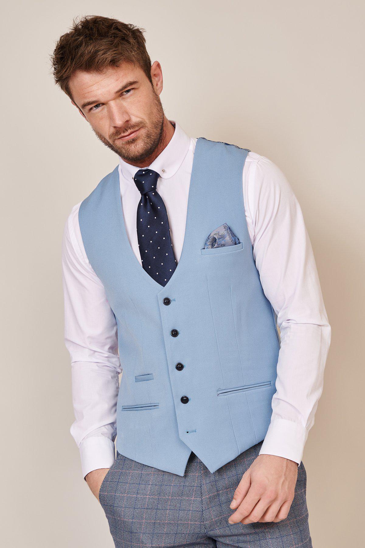A man poses against a neutral background wearing the marcdarcy KELLY Sky Blue Single Breasted Waistcoat with a white dress shirt, navy polka dot tie, plaid trousers, and a pocket square in his vest pocket.