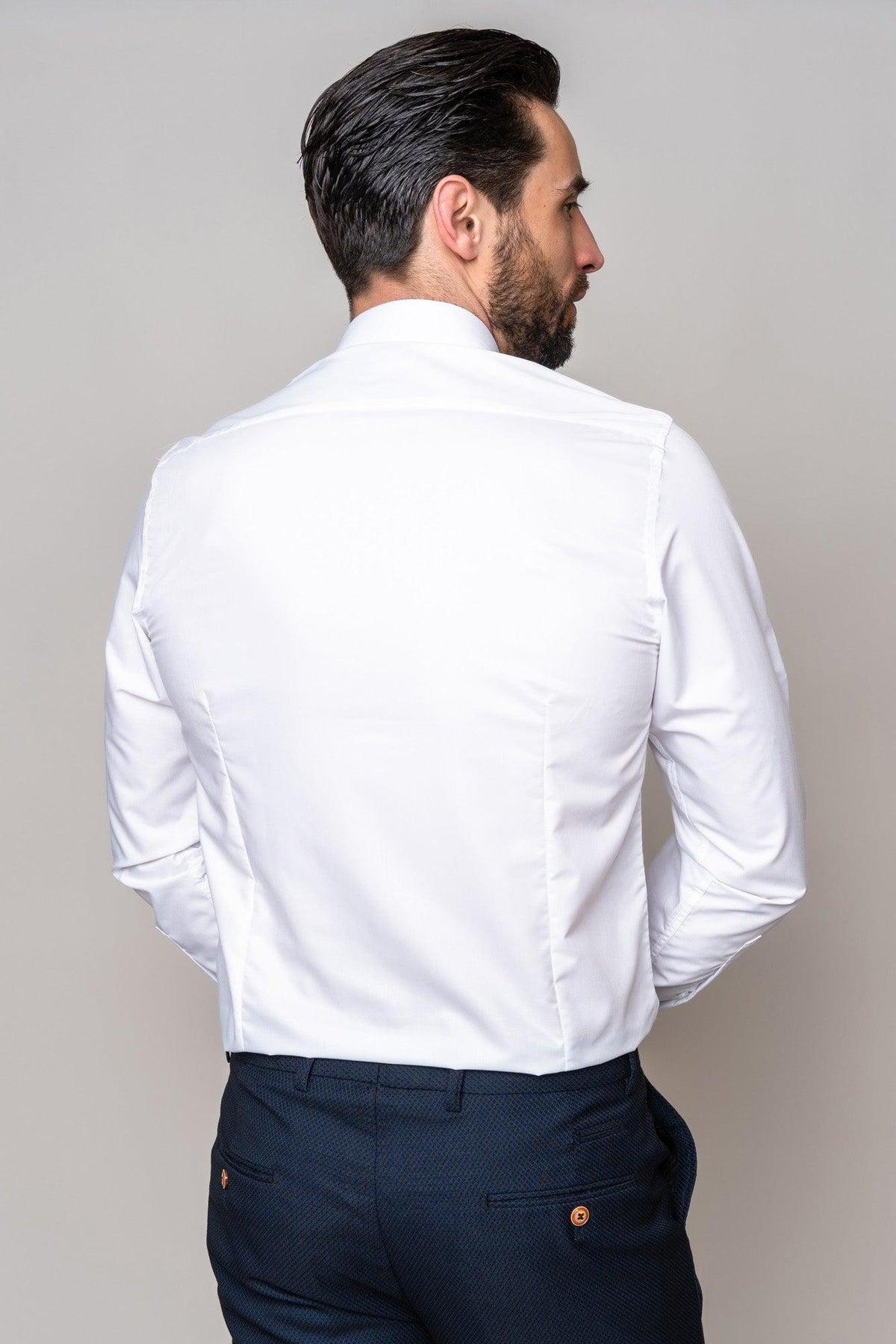 A man with dark hair and a beard stands with his back to the camera, wearing the Marc Darcy CARTER White Long Sleeve Shirt and dark blue trousers. His hands are behind him against a light, uncluttered background.