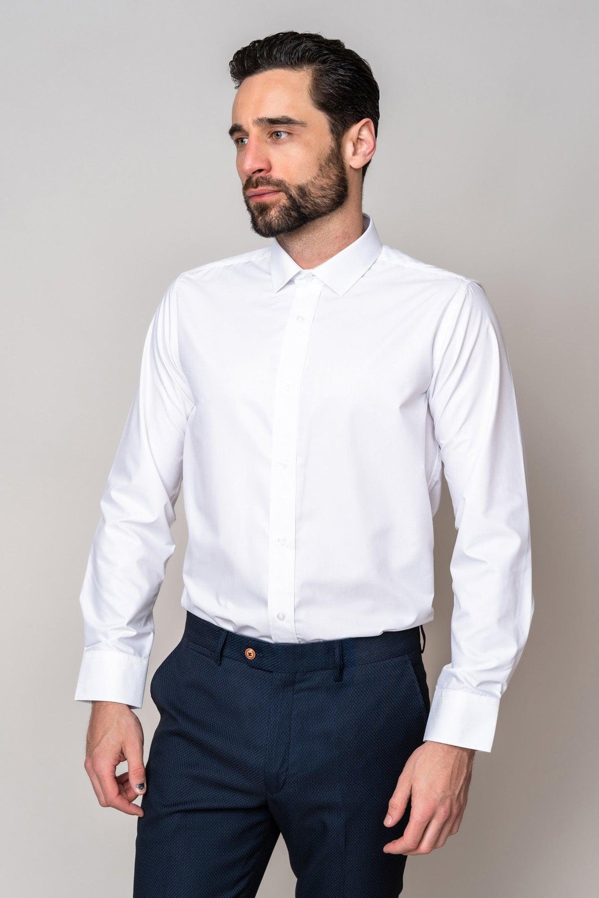A man with dark hair and a beard wears the Marc Darcy CARTER White Long Sleeve Shirt and navy blue pants, standing against a plain light grey background, looking to the side.
