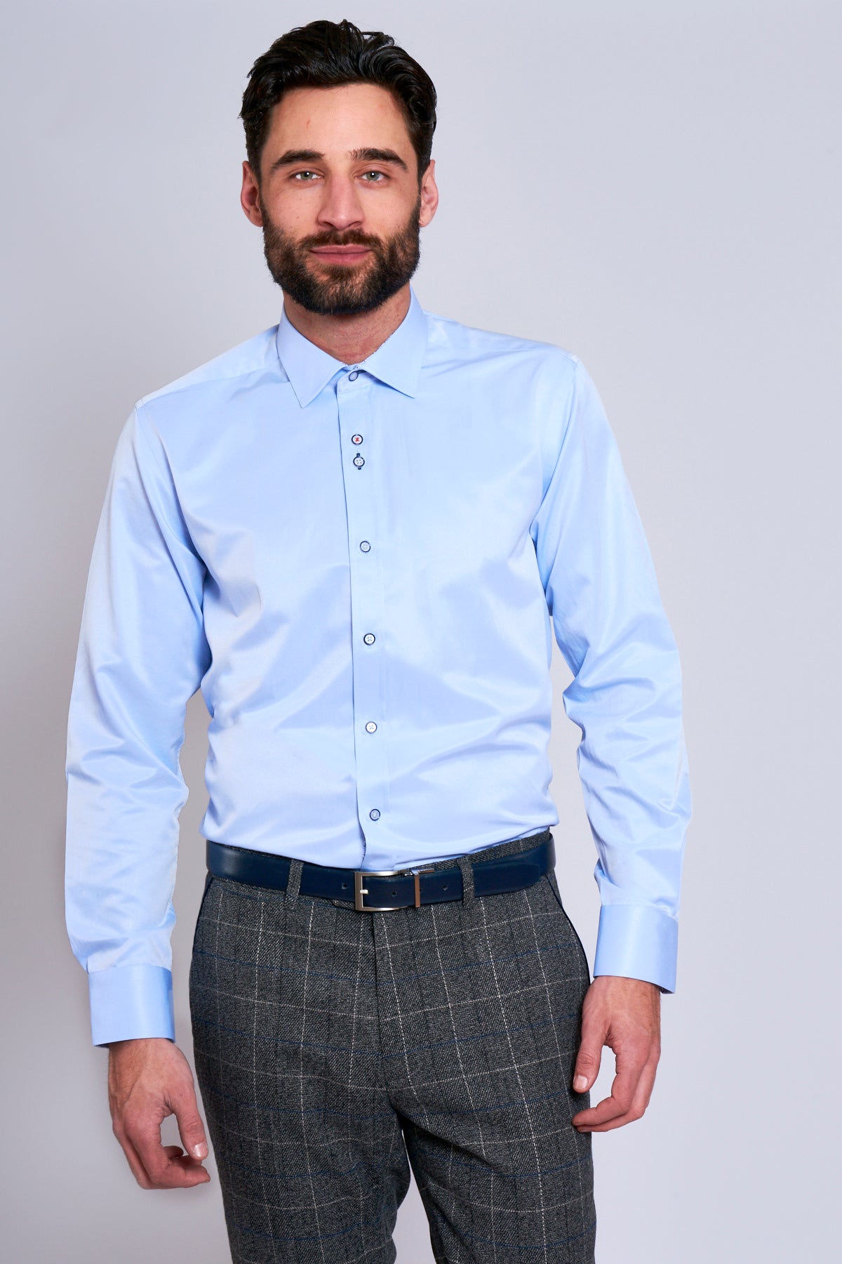 A man with a beard wears the ALFIE - Sky Blue Long Sleeve Shirt by Marc Darcy, paired with grey checked trousers and a dark blue belt for a smart office look.