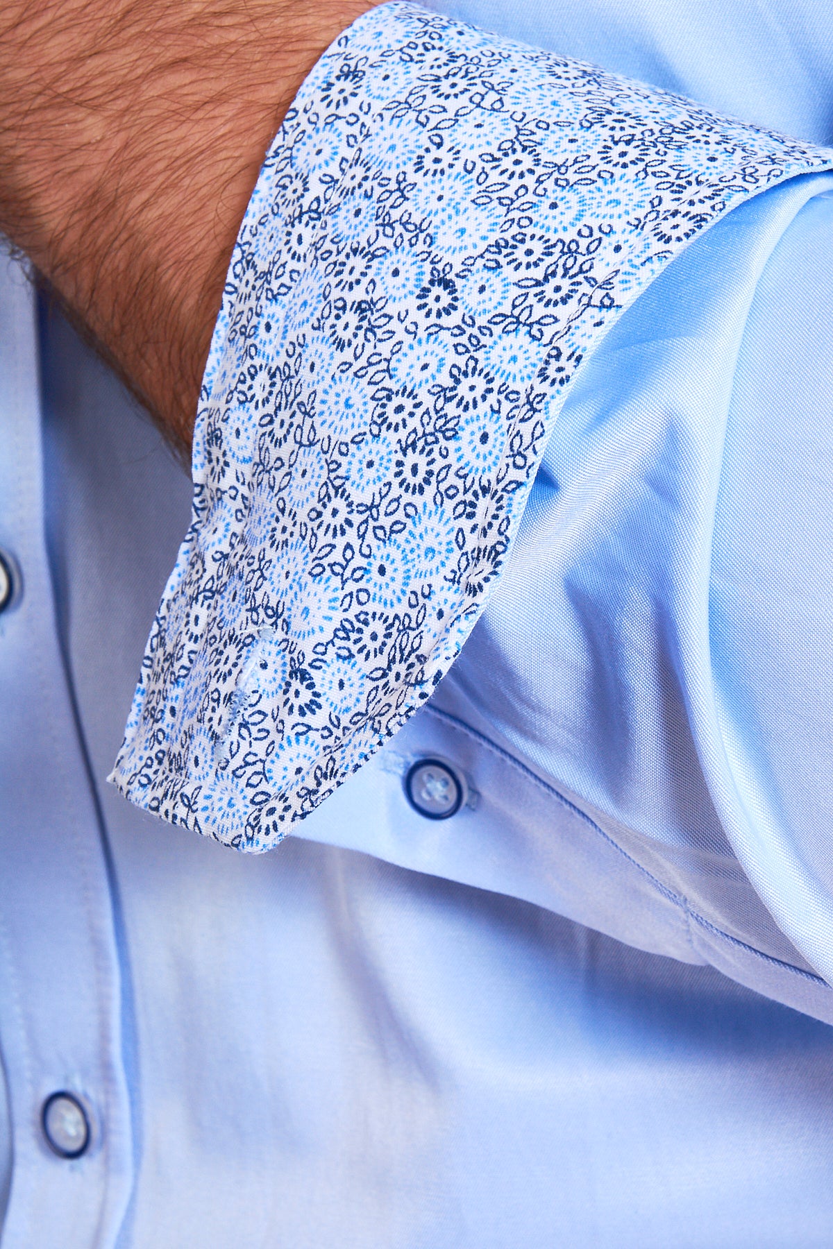 A close-up of a person’s arm in the Marc Darcy ALFIE Sky Blue Long Sleeve Shirt reveals a detailed blue and white floral cuff lining. Made from a cotton blend with light blue buttons, it's perfect for refined office attire.